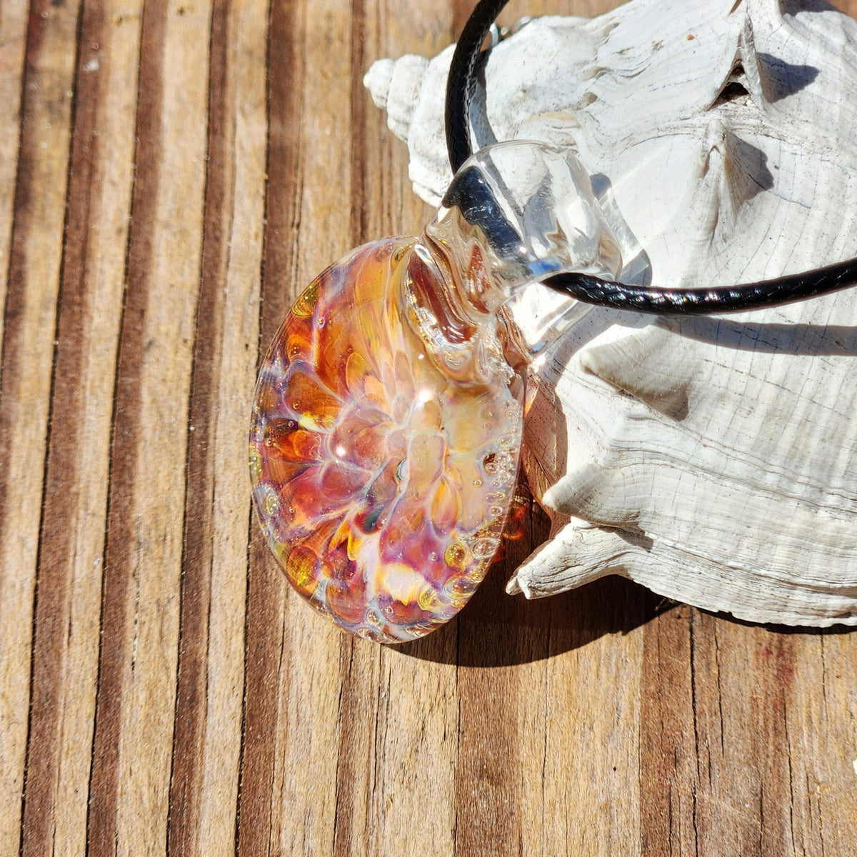 Side view of the glass sunset pendant necklace, highlighting the clear glass and vivid swirling colors, on a white shell background.