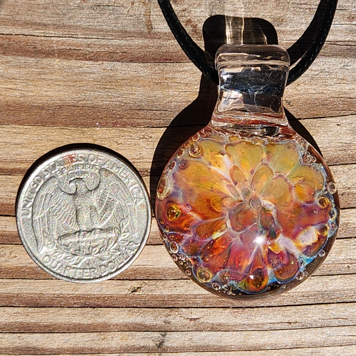Jewelry close-up featuring the glass sunset pendant with intricate color swirls, worn on a black cord, held against a person's hand.