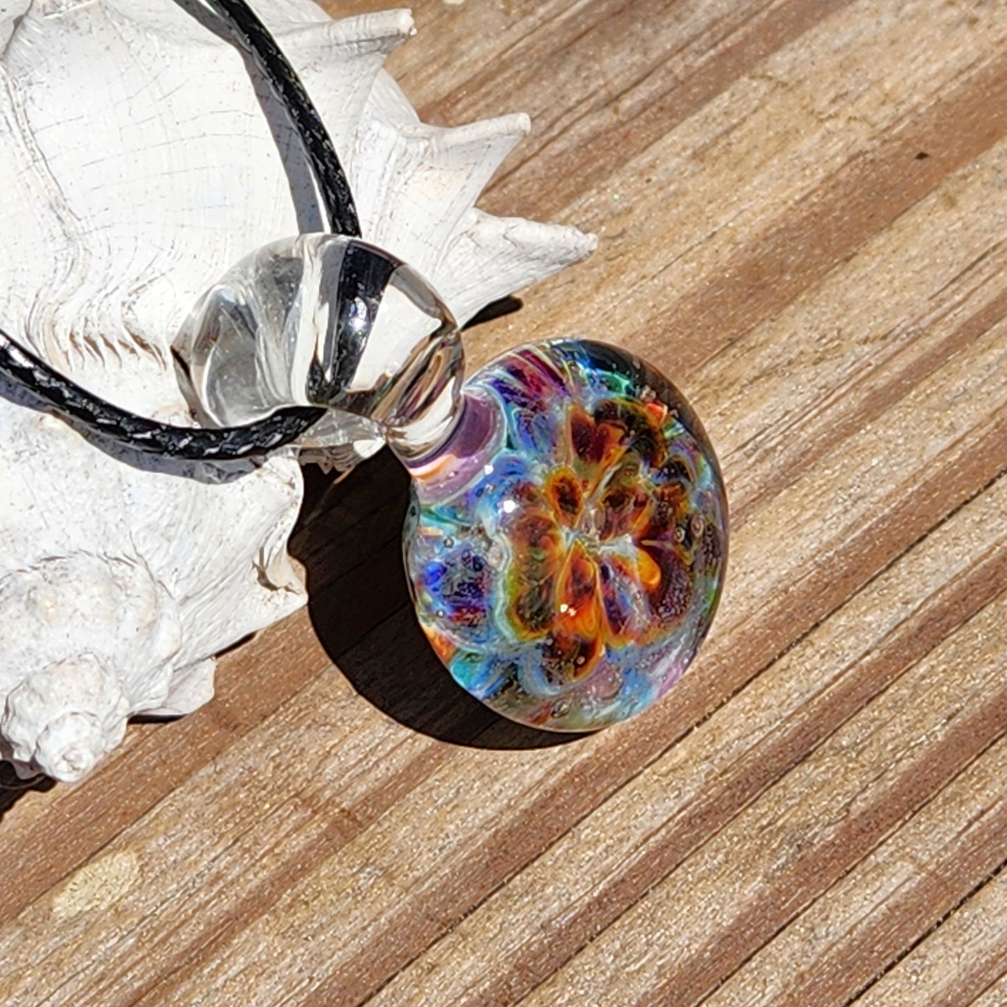 Side view of the colorful glass pendant showing its round shape and detailed swirling pattern, on a sandy surface, SKU: Maly 116