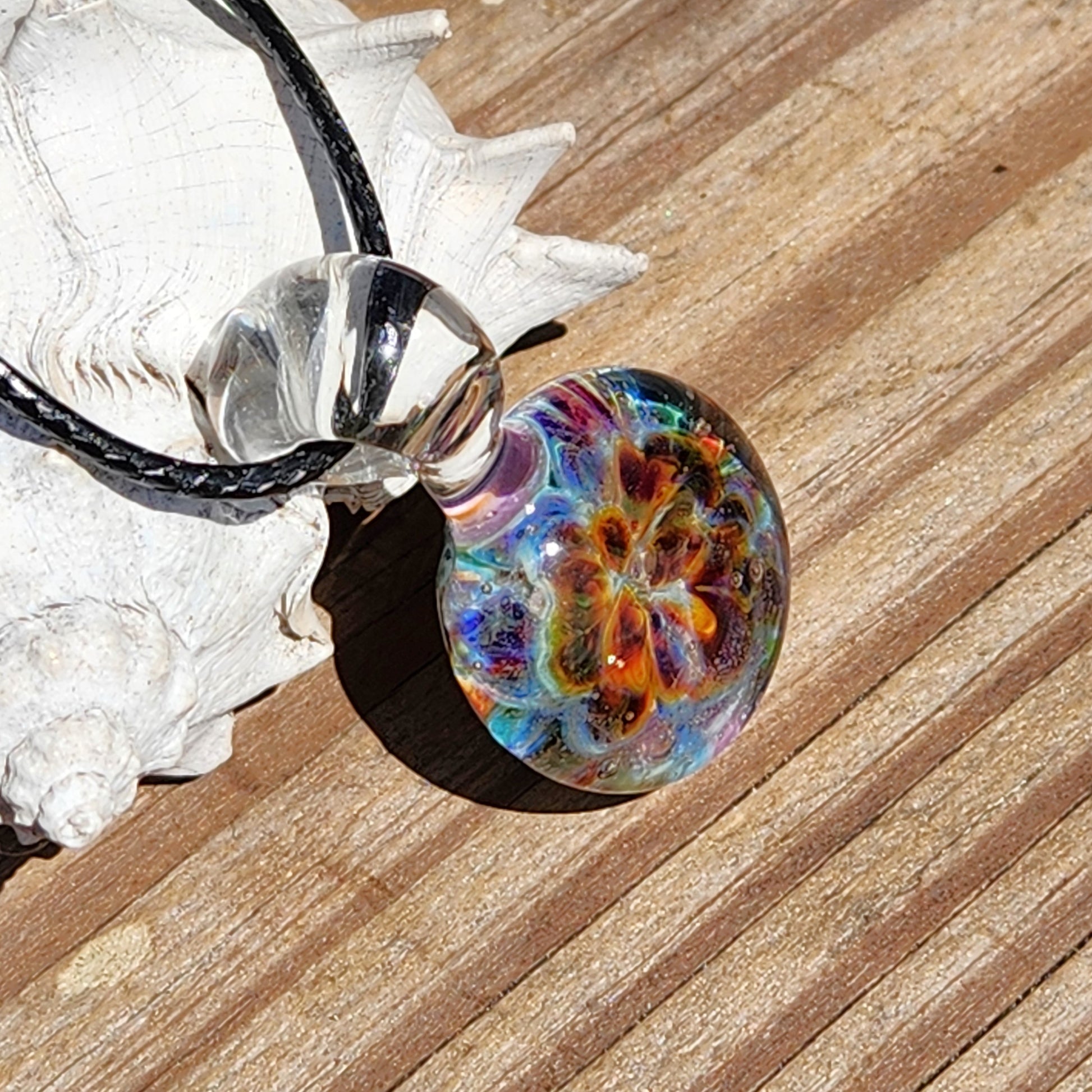 Side view of the colorful glass pendant showing its round shape and detailed swirling pattern, on a sandy surface, SKU: Maly 116