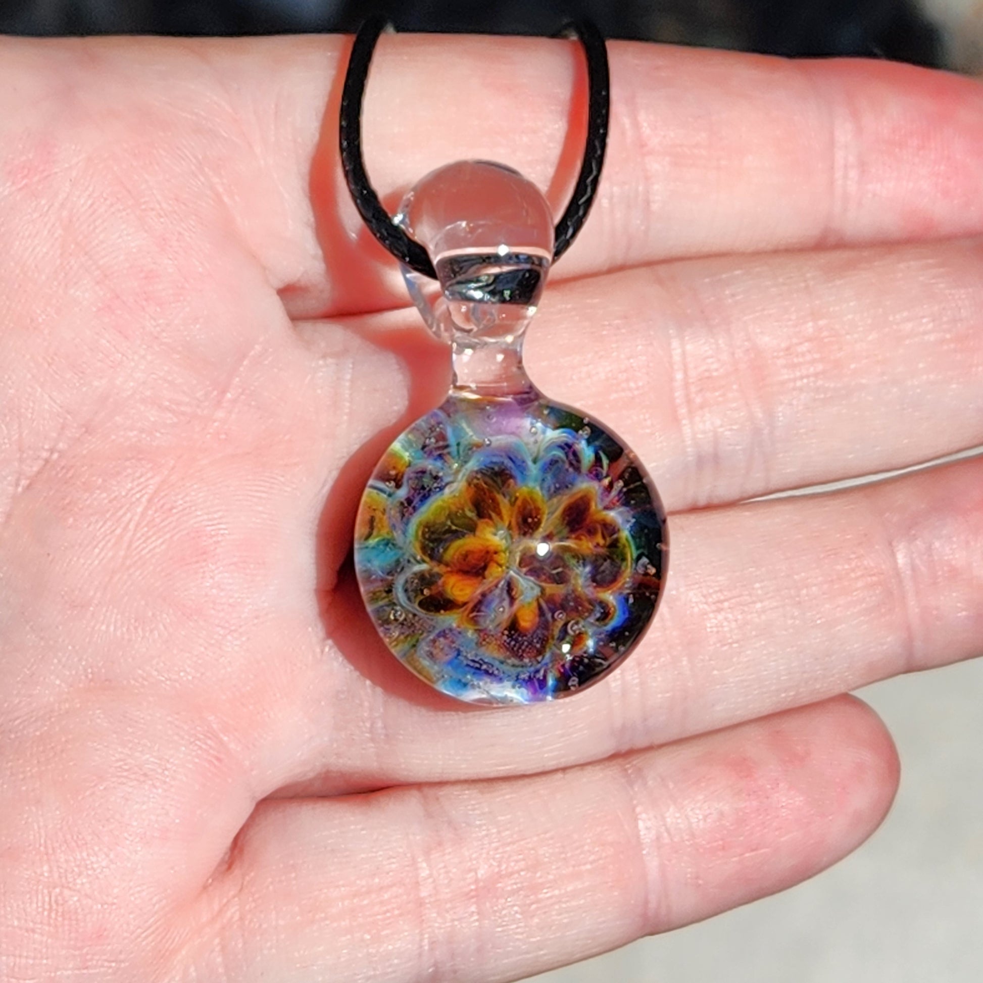 Colorful glass pendant with swirling patterns in vibrant hues, attached to a black cord, worn on a finger for jewelry display