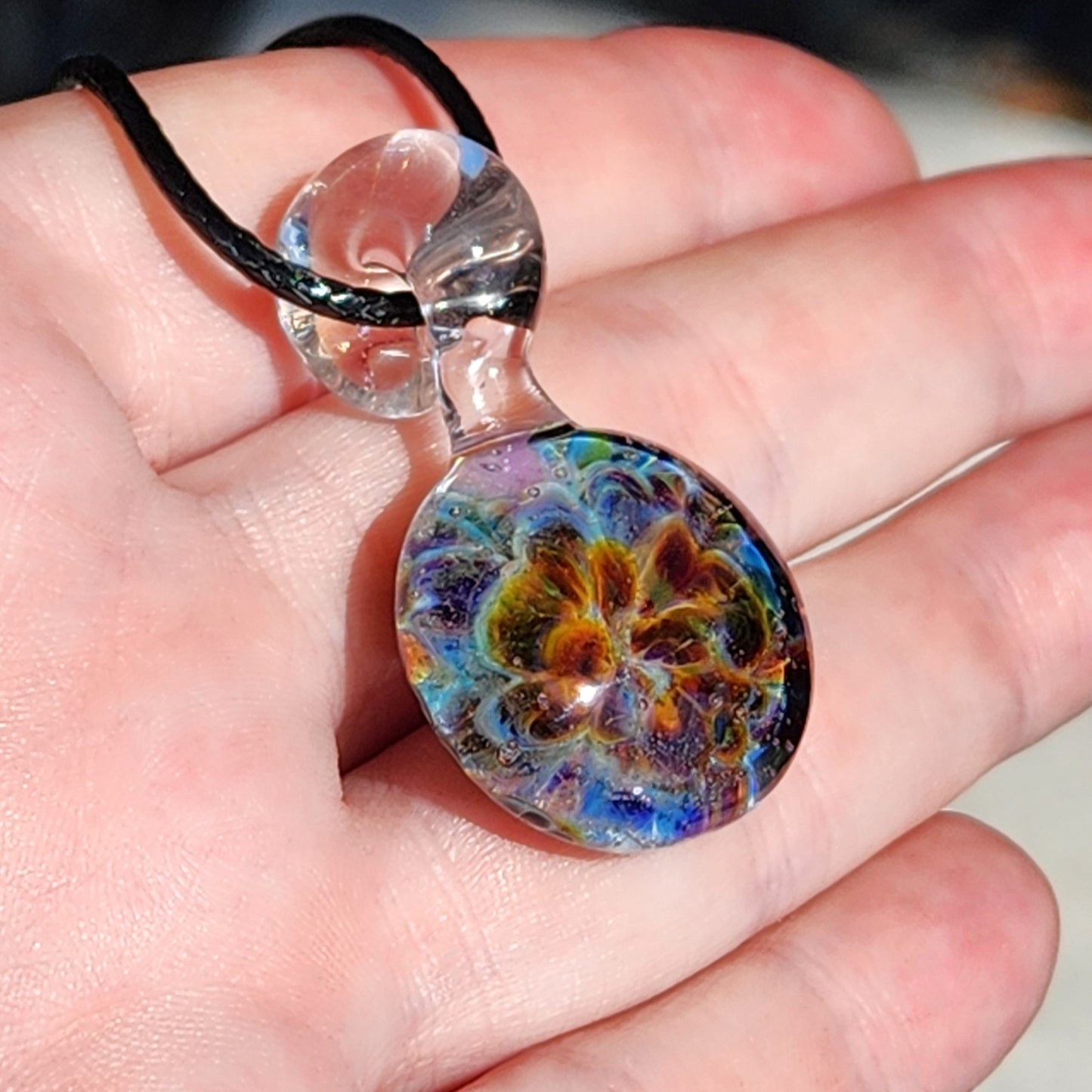 Close-up of a handmade glass pendant with vibrant swirling colors, attached to a black cord, resting on a textured wooden surface.