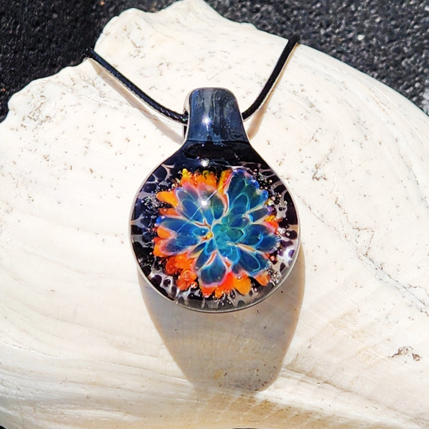 Handmade glass pendant necklace featuring a vibrant blue flower design with orange accents, black cord, on a sandy surface
