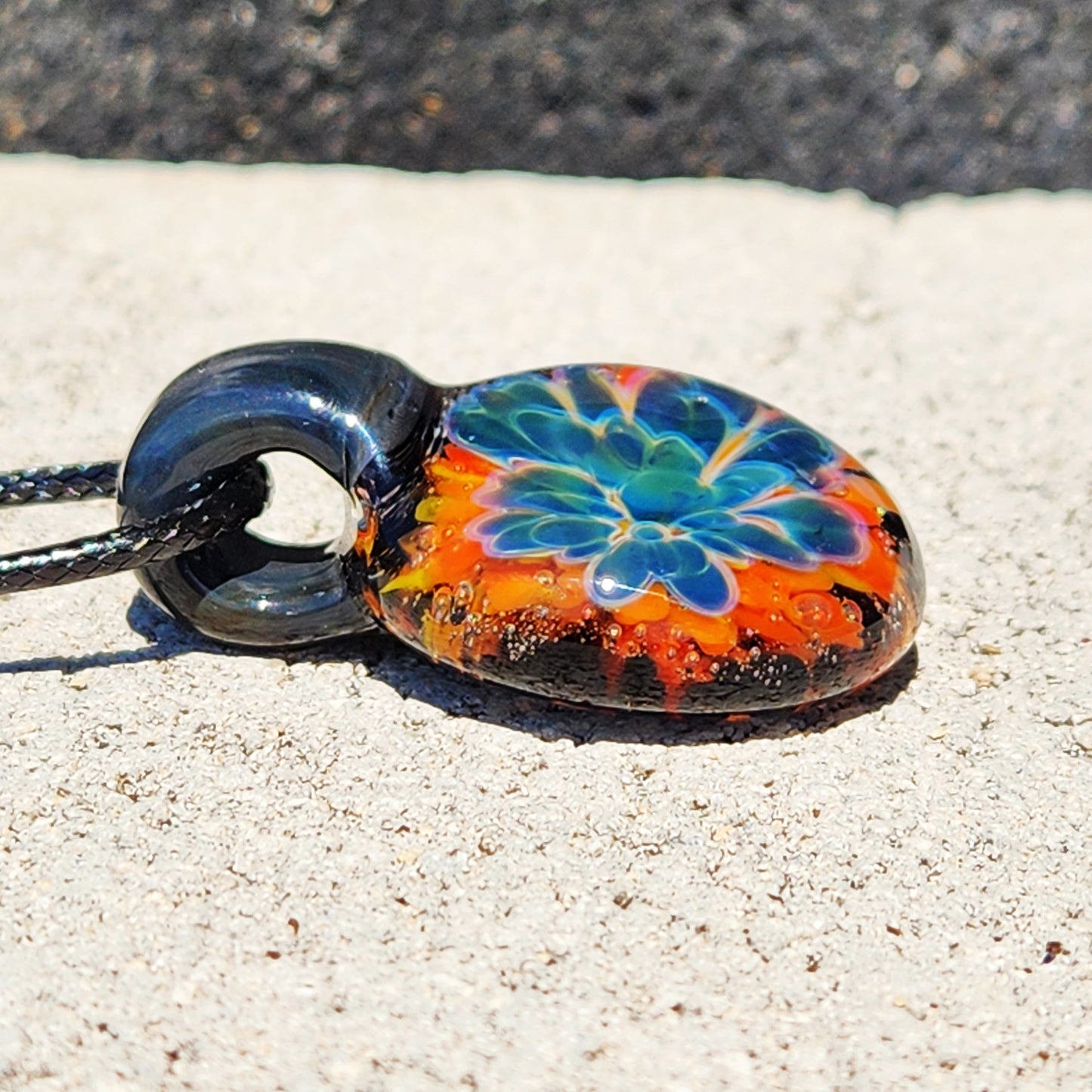 Detail of the handmade glass floral pendant with bright colors, black cord, resting on a sandy surface