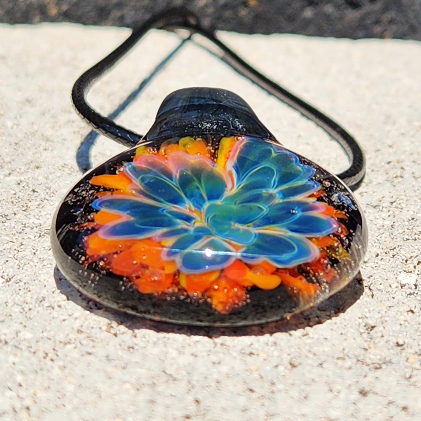 Top view of the colorful glass pendant with floral design, black cord, on a sandy background