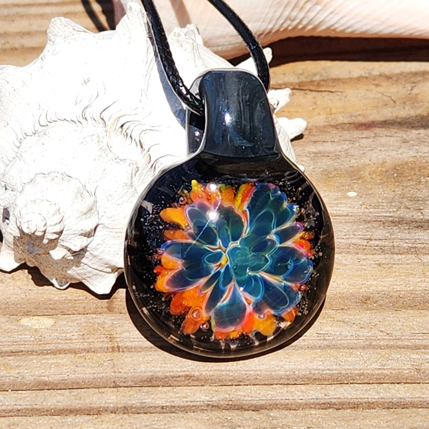 Handmade glass pendant necklace featuring a vibrant blue flower design with orange accents, black cord, on a wooden surface.
