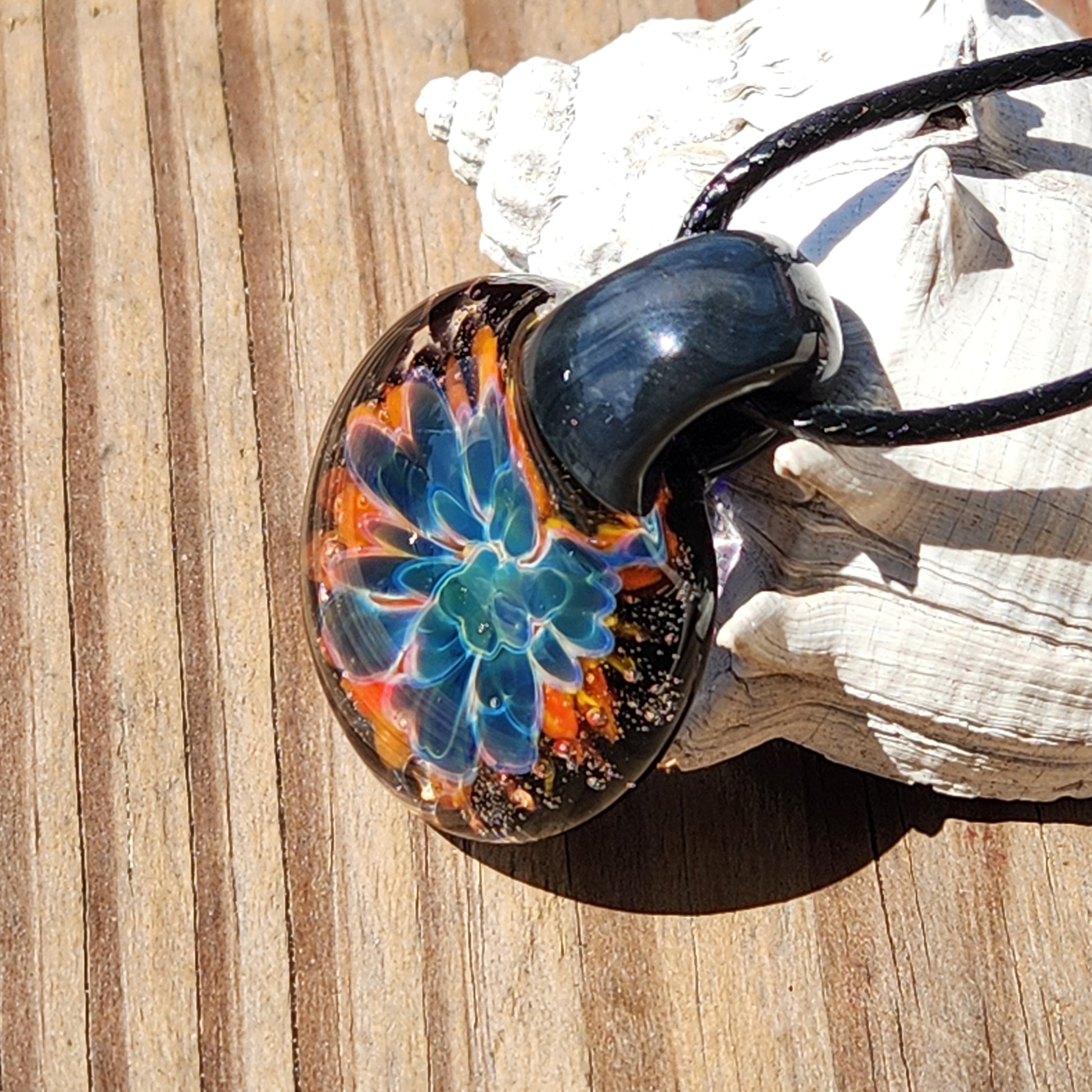 Side view of the vibrant glass flower pendant with black cord, on a sandy surface