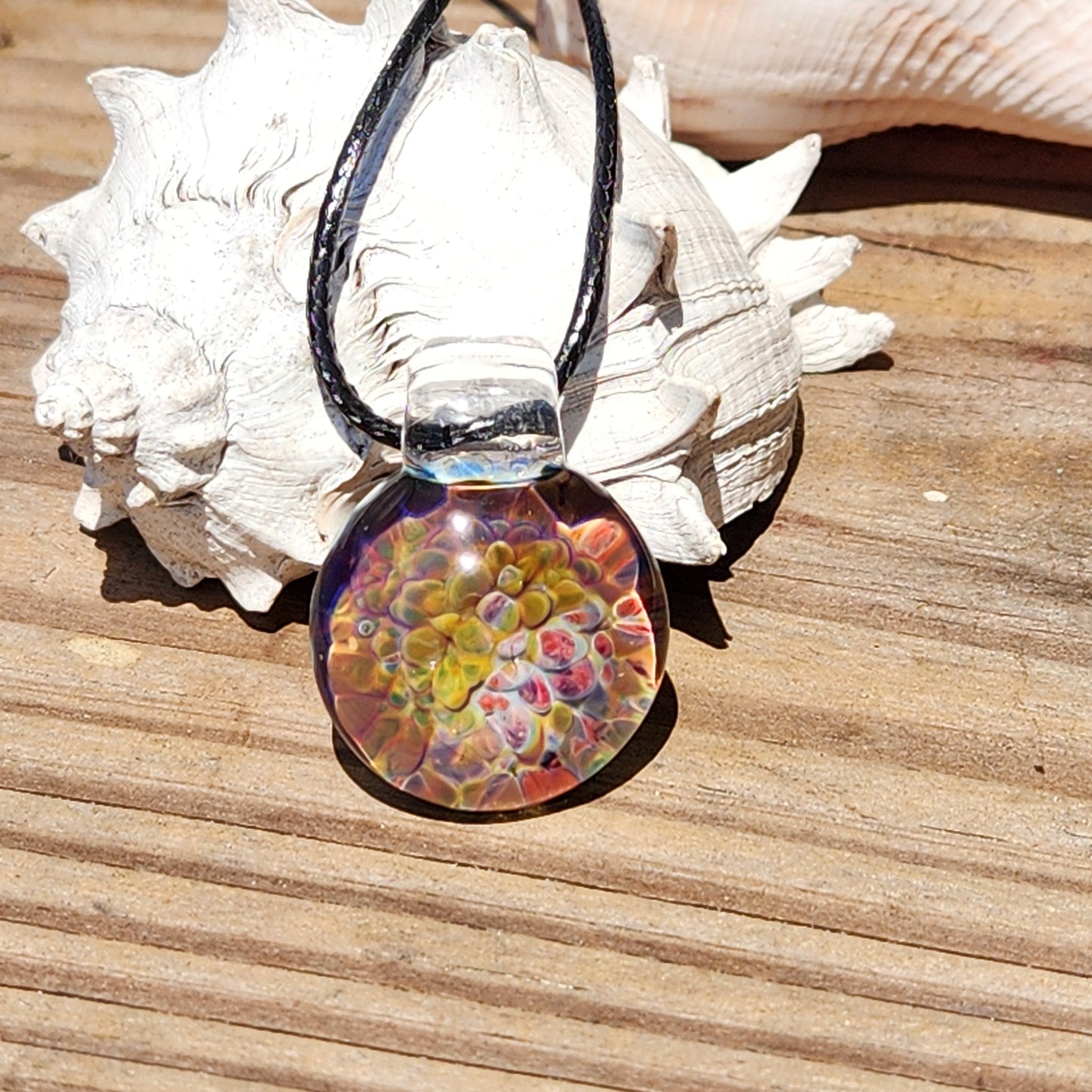 Detailed shot of the glass pendant's vibrant, swirling colors, placed on a textured wooden surface with shell accents.