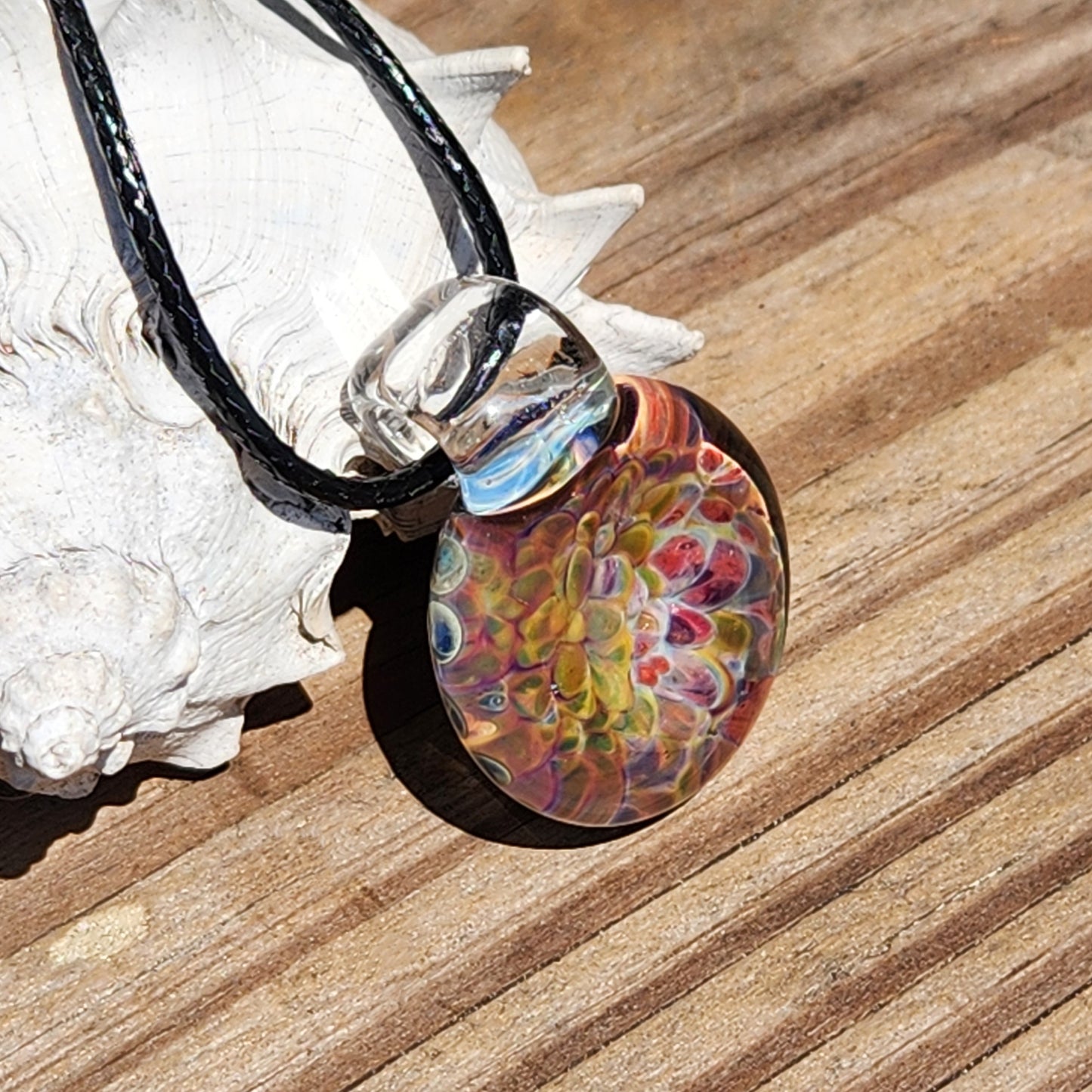 Close-up of the glass pendant showing intricate multicolor patterns, resting on a white shell with a textured surface.