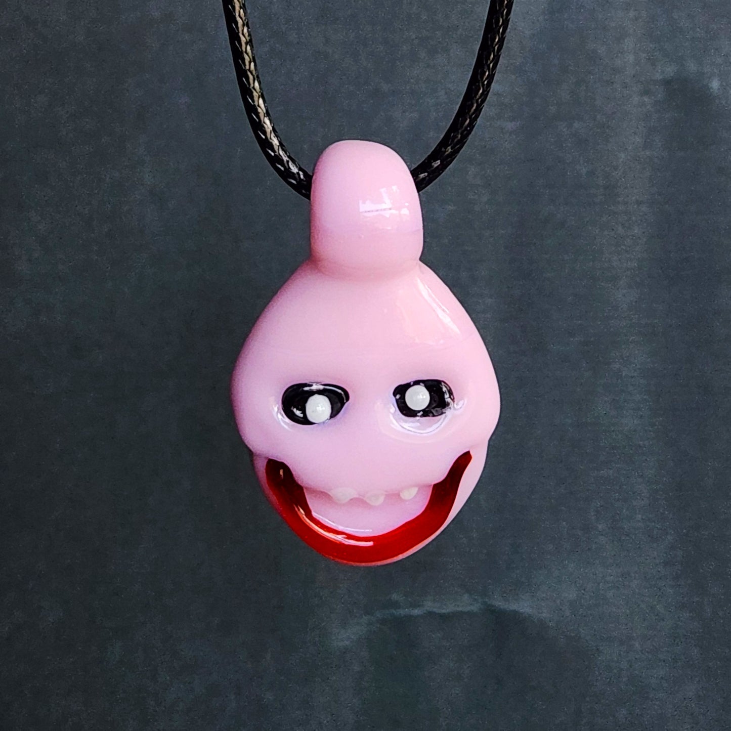 A pink novelty pendant necklace featuring a stylized cartoon face with black and white eyes and a wide red grin, suspended from a black cord against a dark gray background.