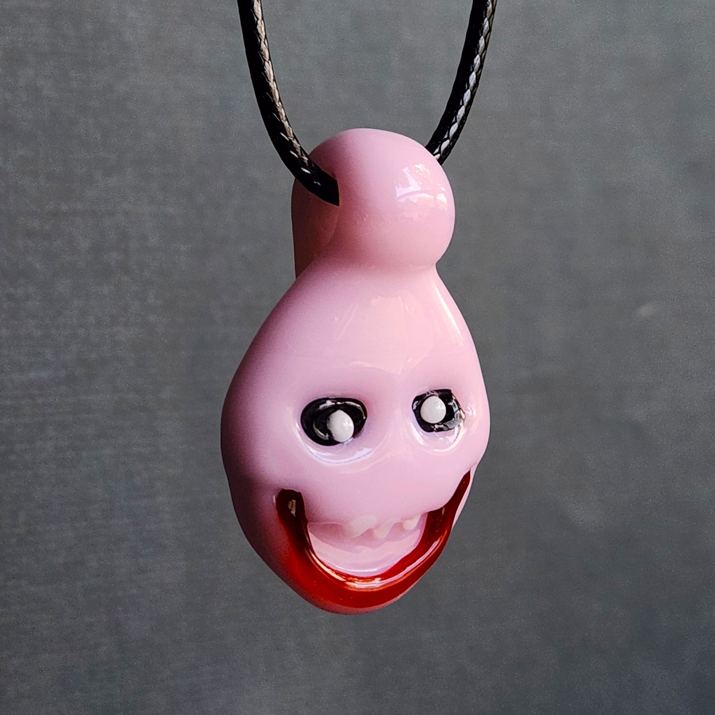 A glossy pink novelty pendant necklace with a cartoonish face, featuring black and white eyes and a red open mouth, hanging from a black cord against a textured gray background.