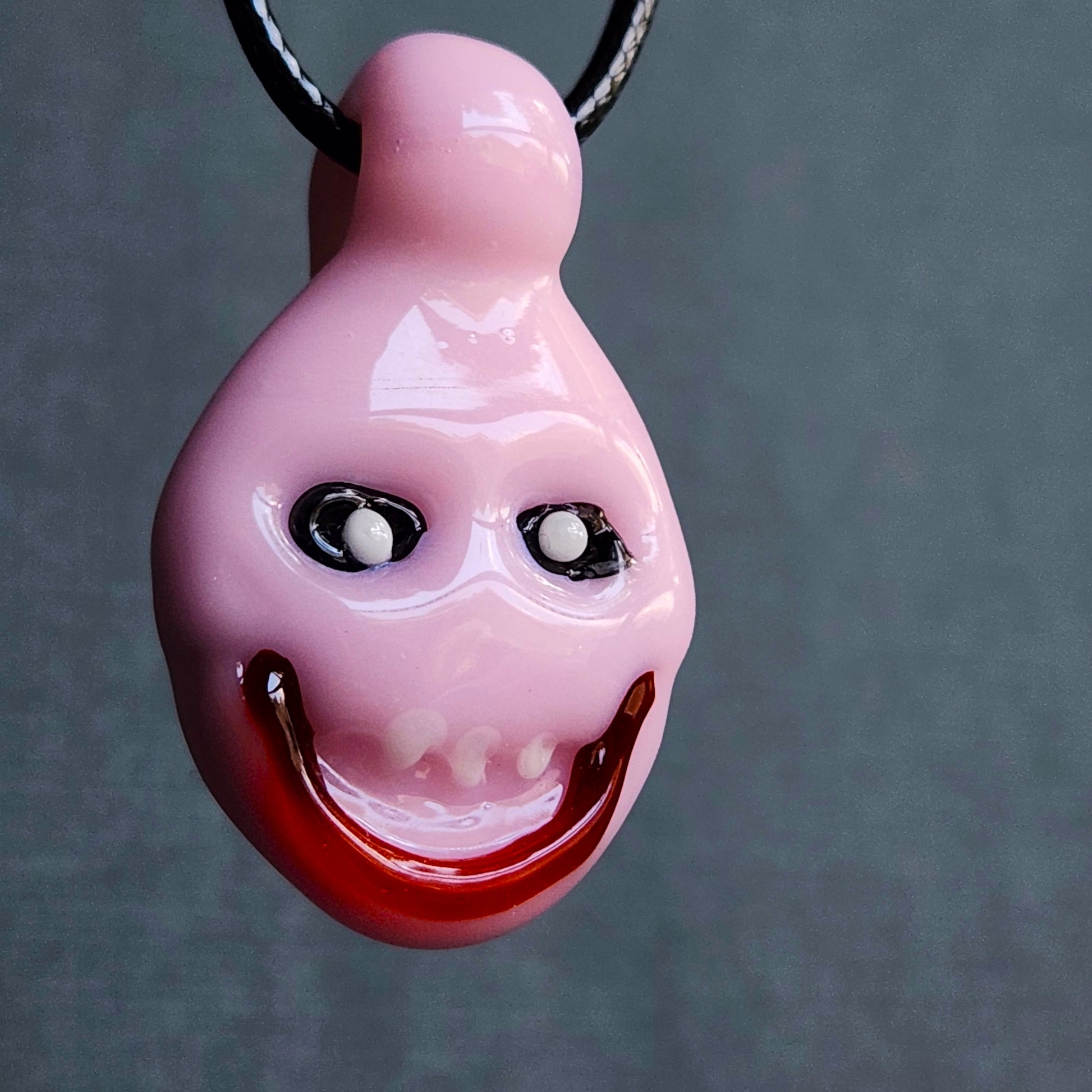A glossy pink teardrop-shaped pendant necklace with a creepy, distorted smile and black-and-white eyes, suspended from a black cord against a dark gray background.