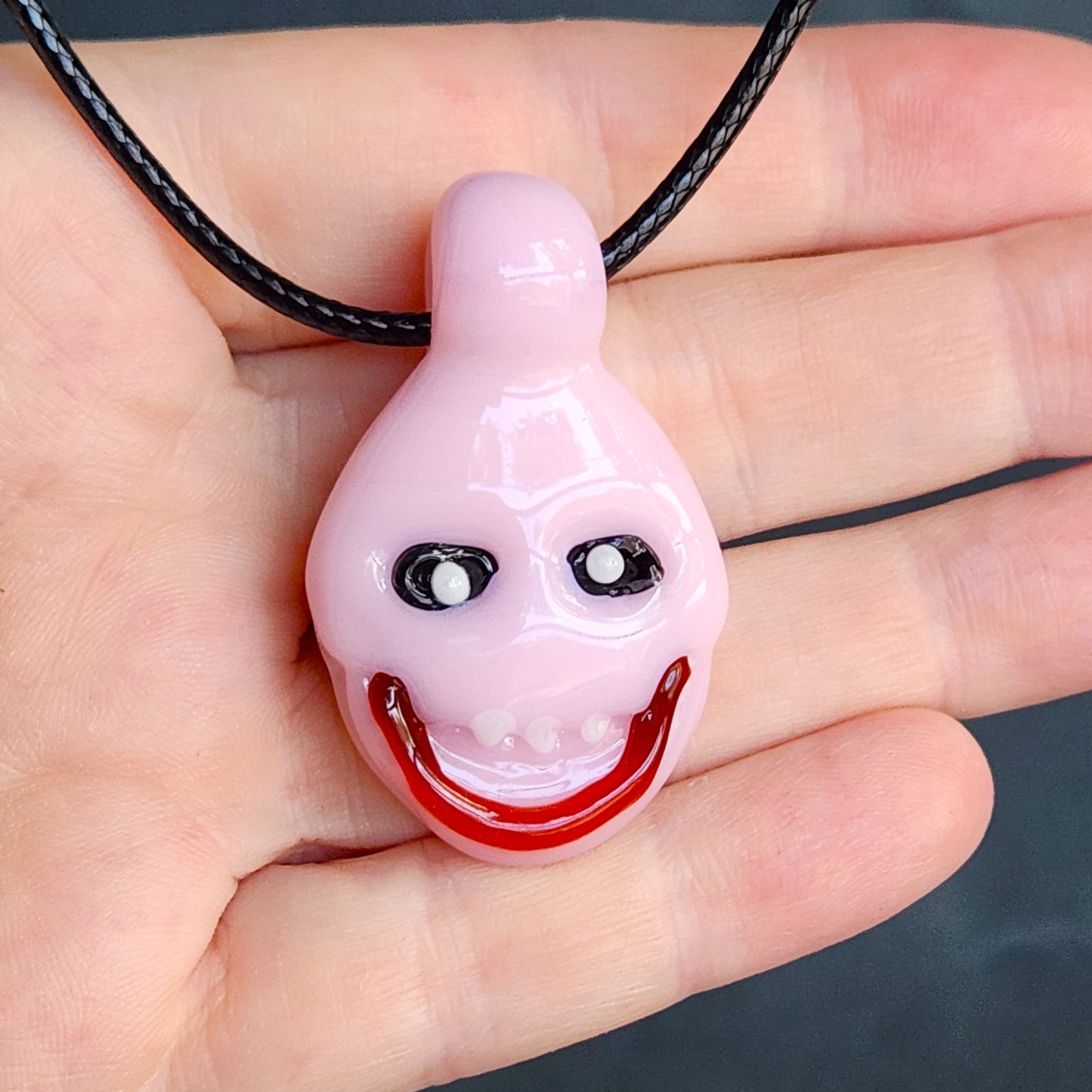 A glossy pink teardrop-shaped pendant necklace with a creepy smiling face, held in a person's hand on a black cord.