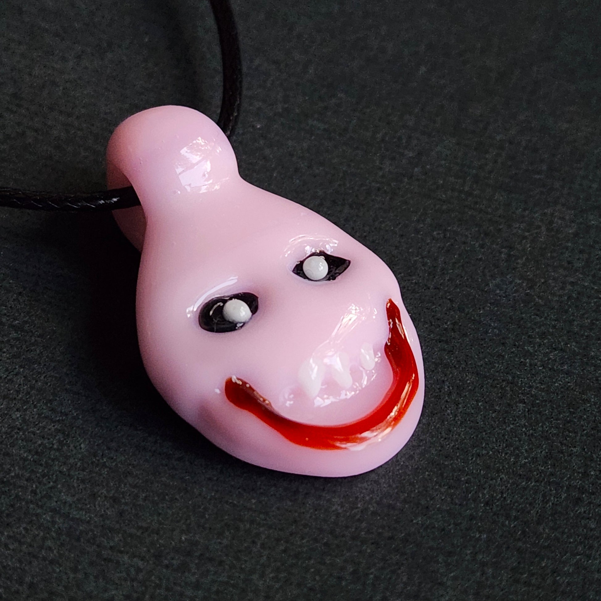 A glossy light pink pendant shaped like a distorted, unsettling face with black and white eyes and a wide red grin, hanging on a black cord against a dark gray textured background.