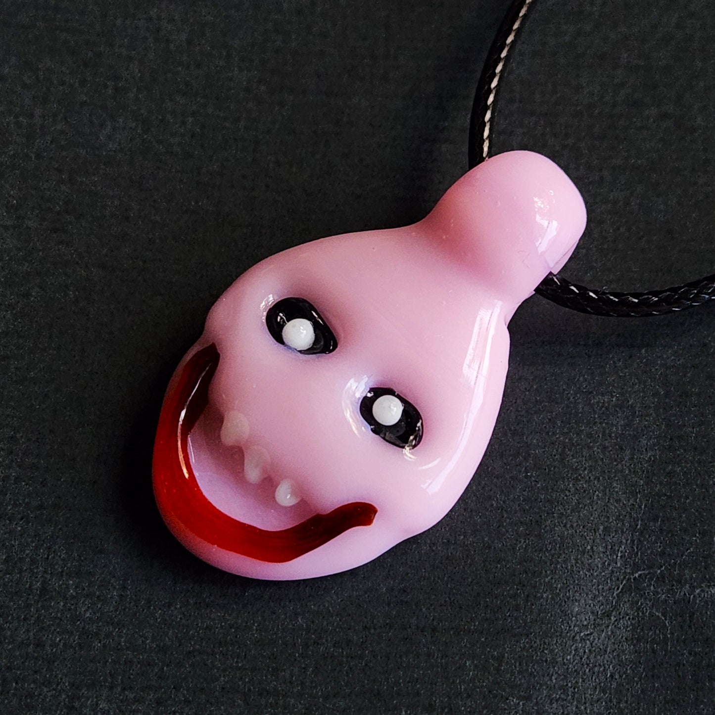 A pink novelty pendant shaped like a distorted cartoon face with large black eyes and an open red mouth, hanging from a black cord on a dark textured background.