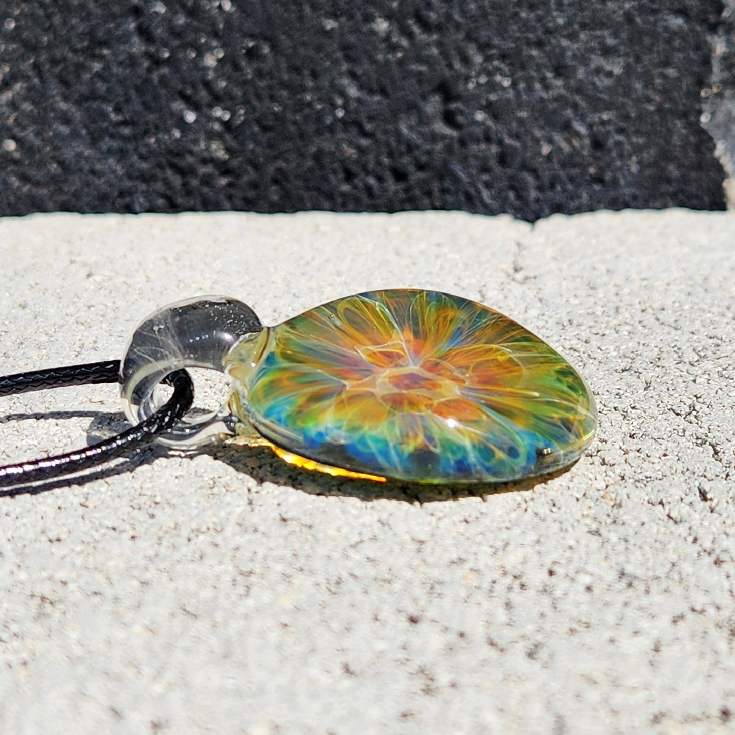 Detailed view of glass pendant with rainbow swirl pattern, black cord, on white shell, sandy background