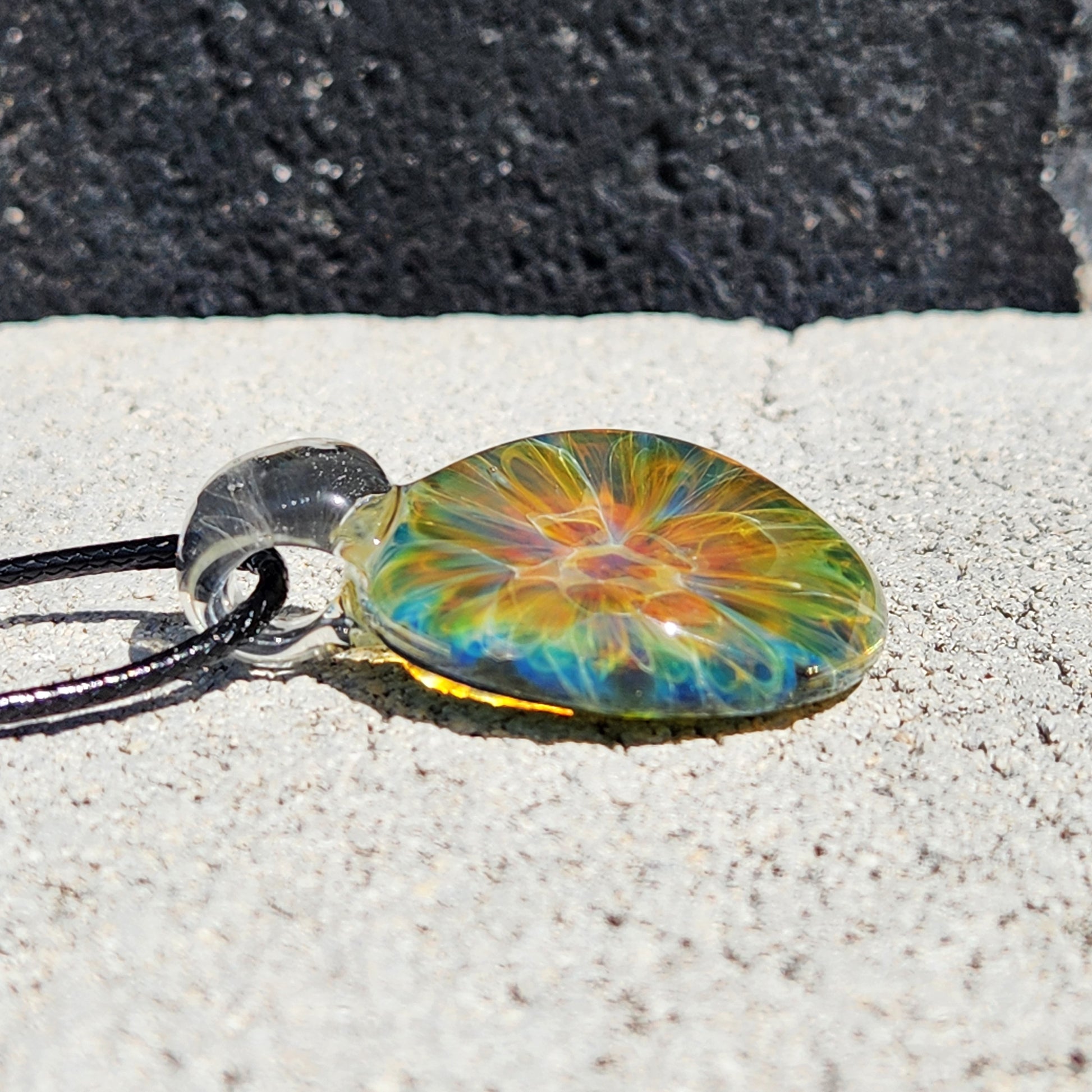 Detailed view of glass pendant with rainbow swirl pattern, black cord, on white shell, sandy background