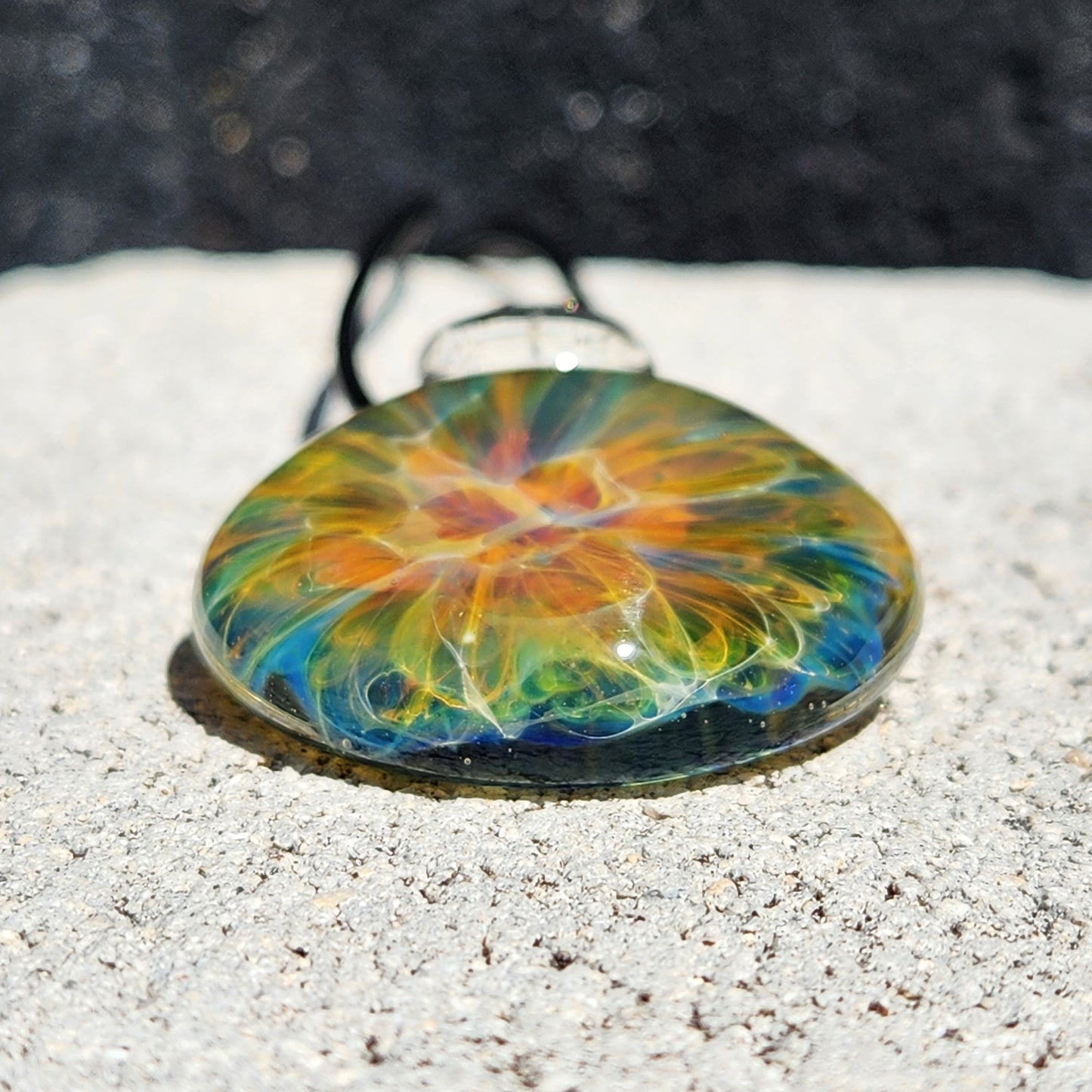 Close-up of glass pendant with rainbow swirl, black cord, on white shell, sandy background