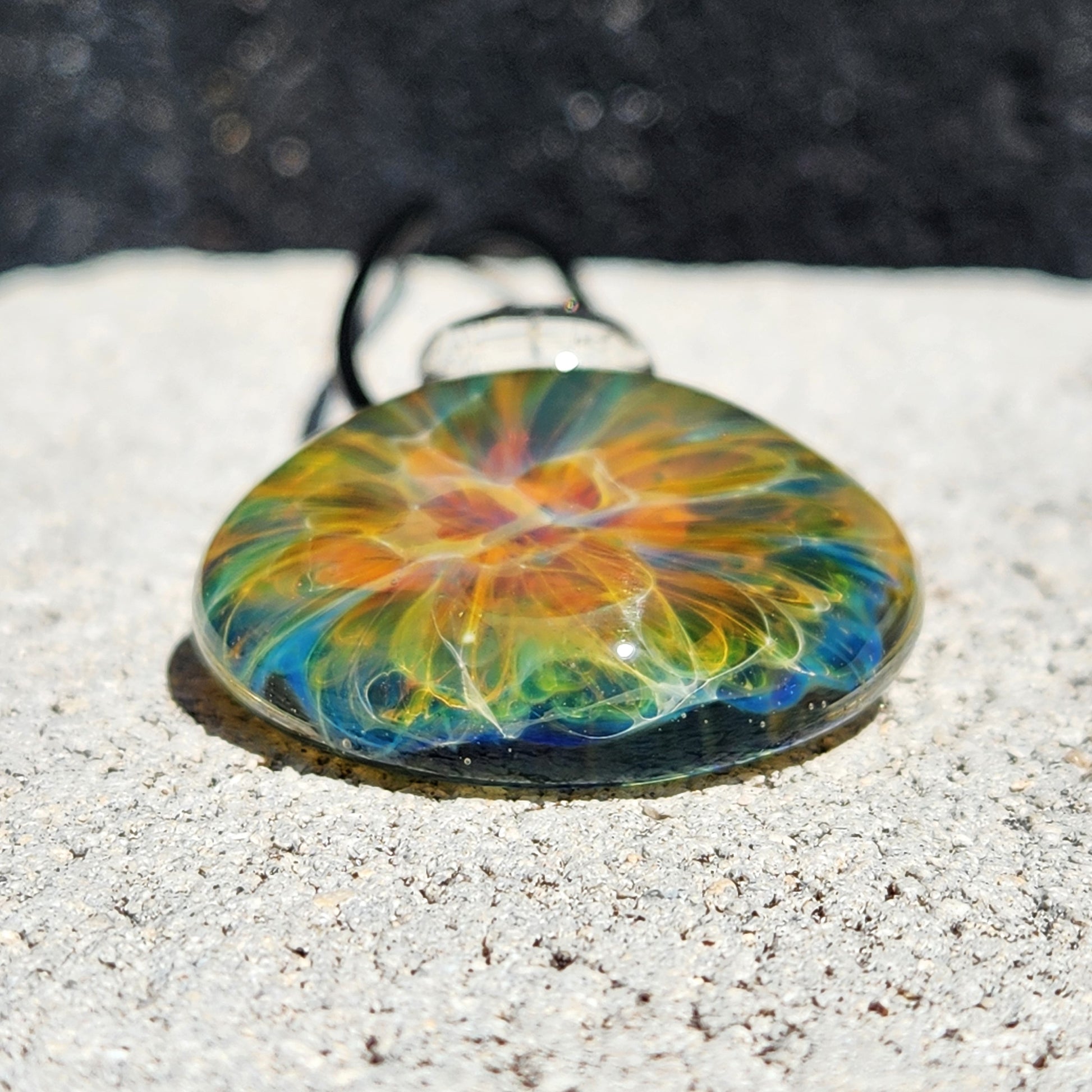 Close-up of glass pendant with rainbow swirl, black cord, on white shell, sandy background