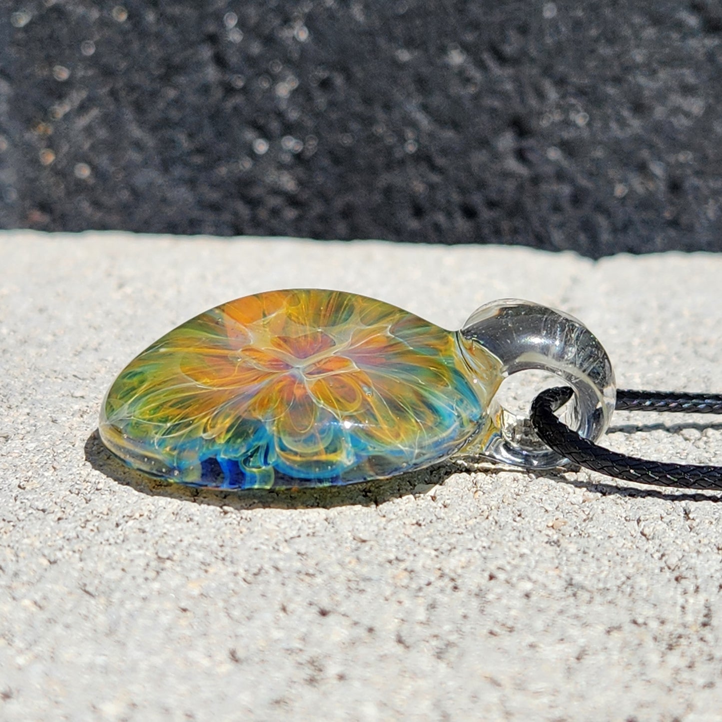Macro shot of the colorful glass pendant showing intricate swirl patterns and reflections, with a black cord and textured background.