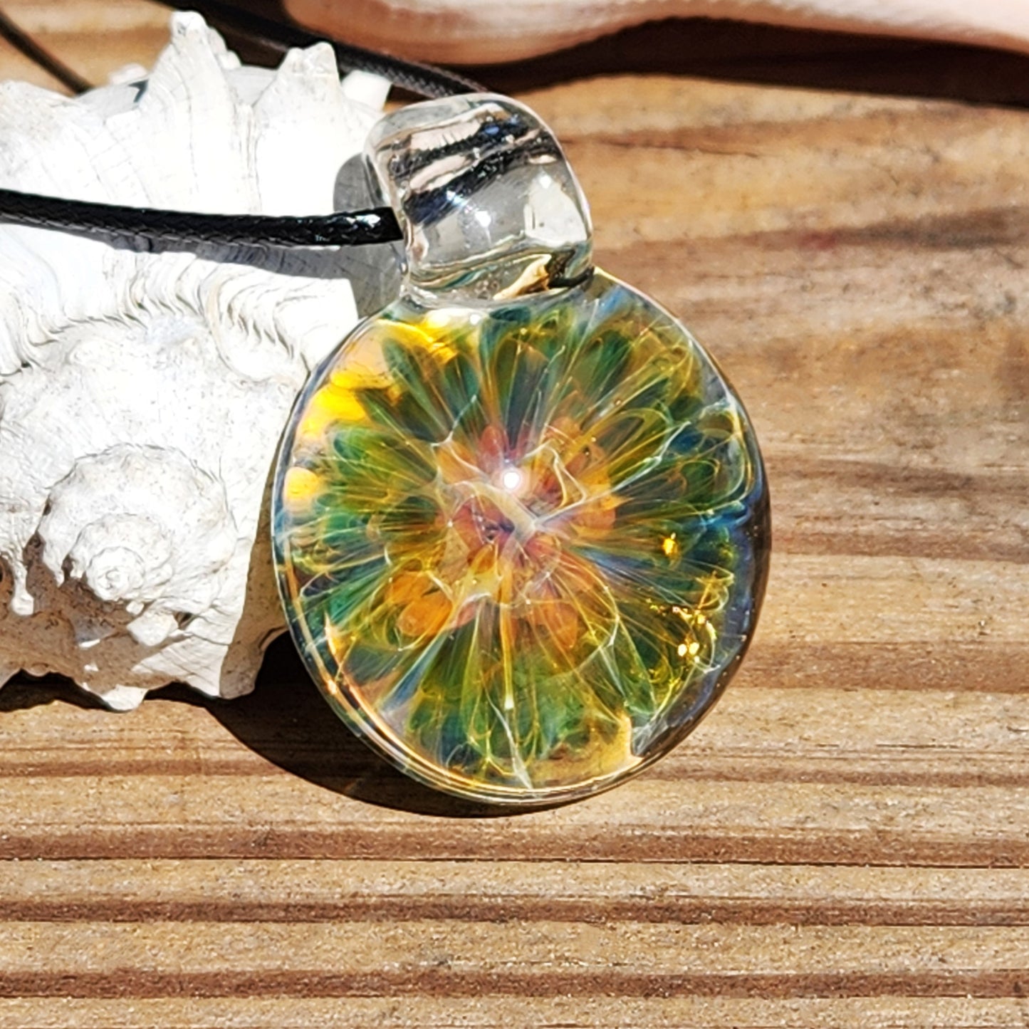 Close-up of a handmade blown glass pendant necklace with vibrant yellow, green, and blue swirls, hanging on a black cord against a wooden background.
