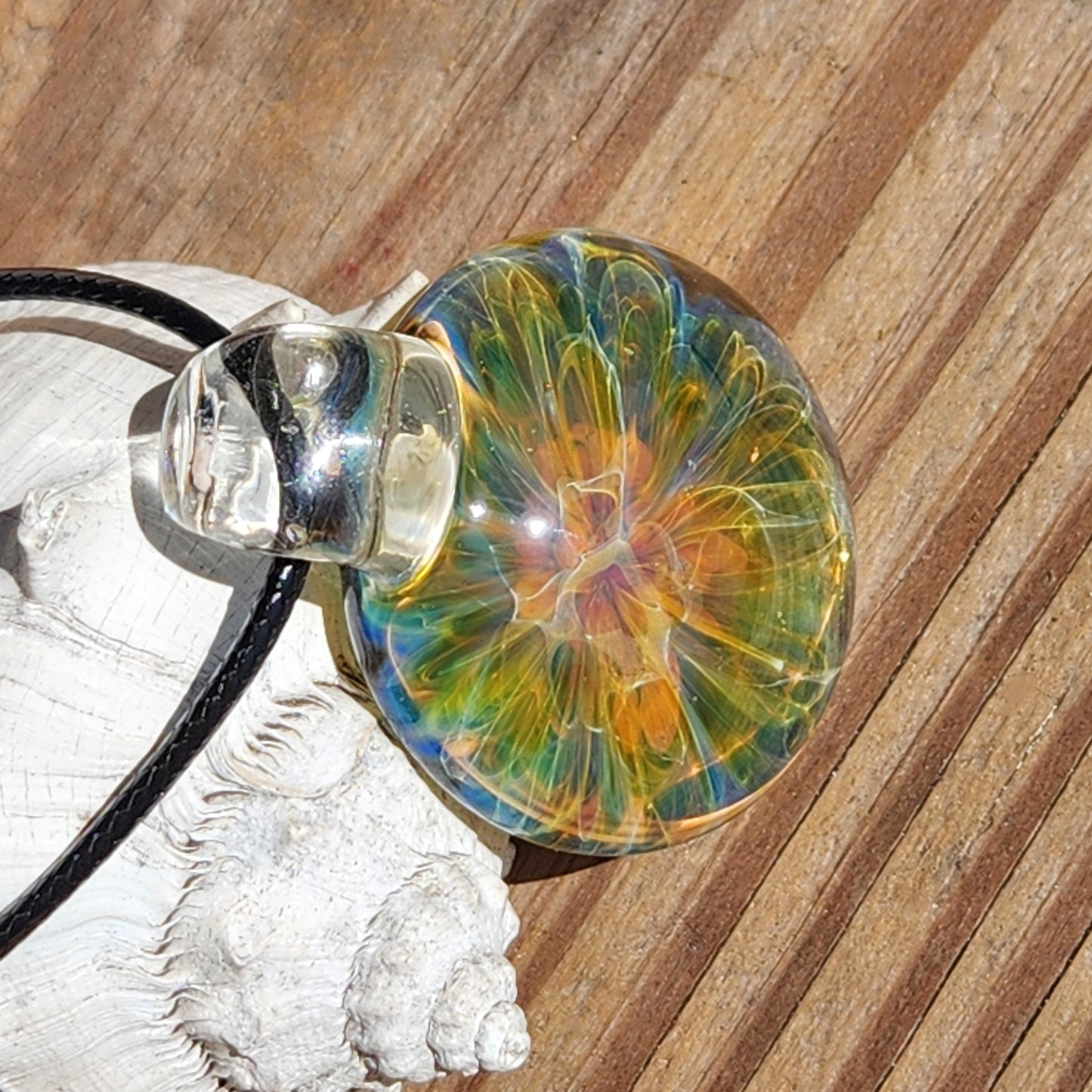 Close-up of colorful glass pendant showing intricate swirl design, black cord, resting on white shell on sandy surface
