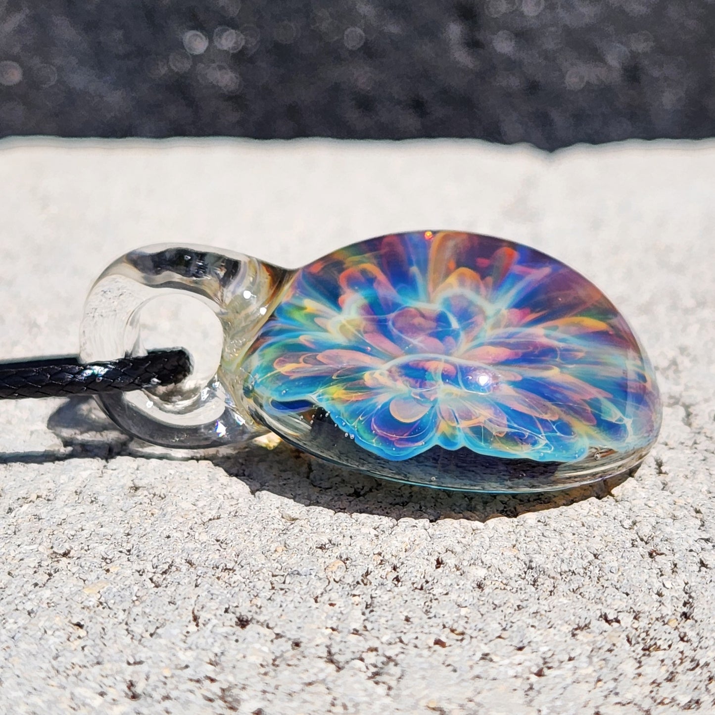 Rainbow glass pendant necklace with intricate swirl pattern, hanging on black cord, photographed on textured white background
