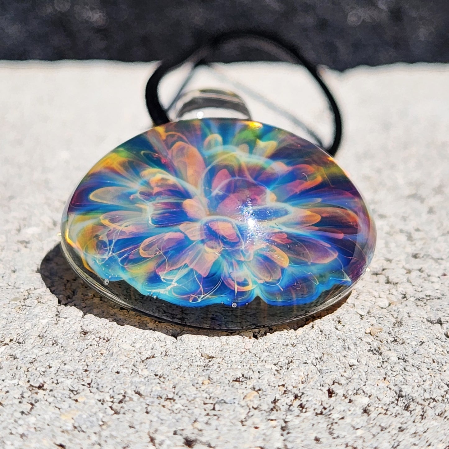 Colorful handmade glass pendant with swirling rainbow hues, round shape, on black braided cord, resting on white textured paper