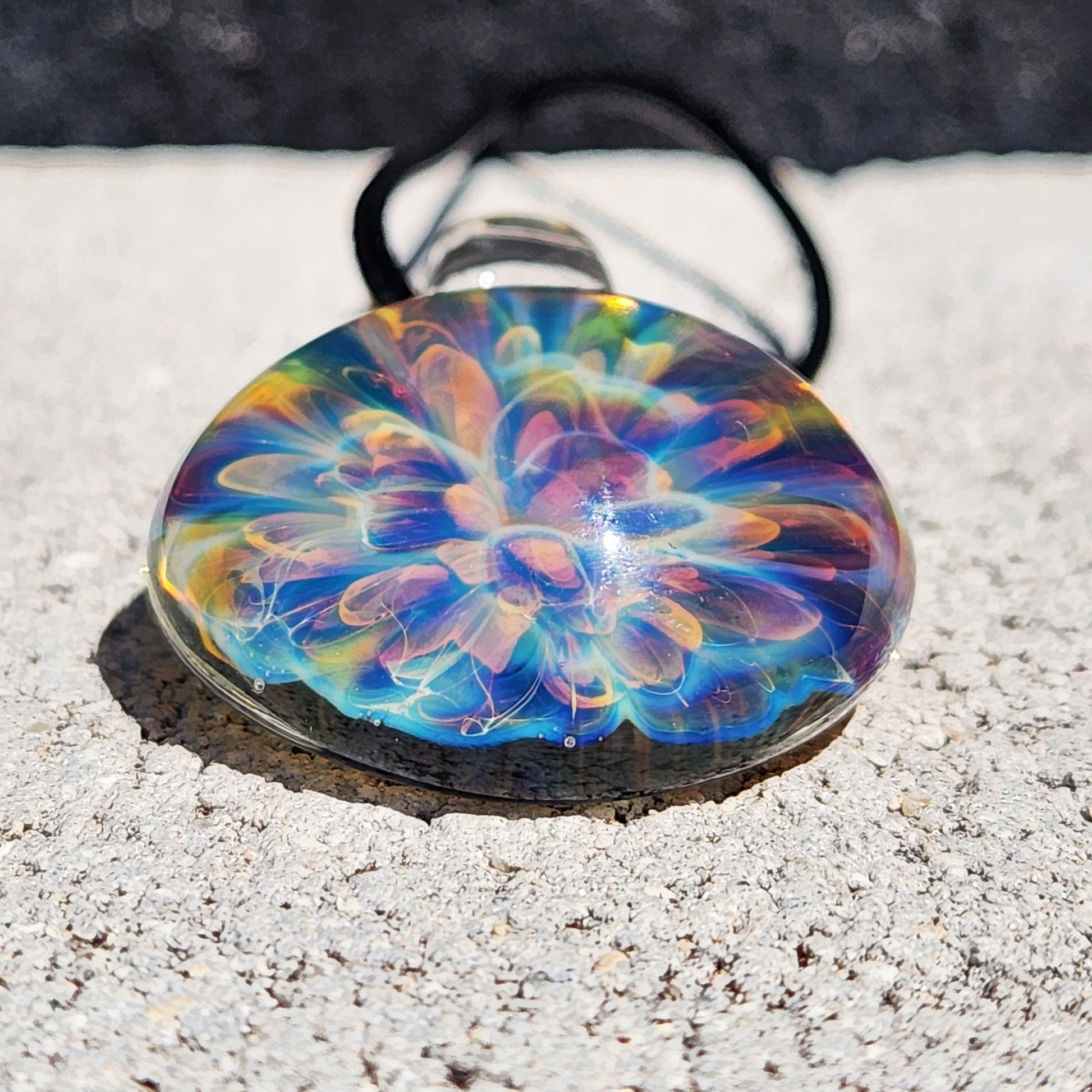Colorful handmade glass pendant with swirling rainbow hues, round shape, on black braided cord, resting on white textured paper