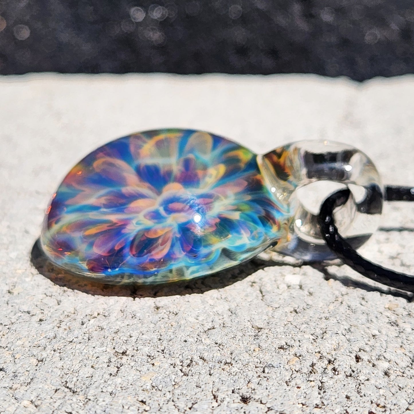Rainbow glass pendant with vibrant swirl pattern, on black cord, placed on white textured paper surface