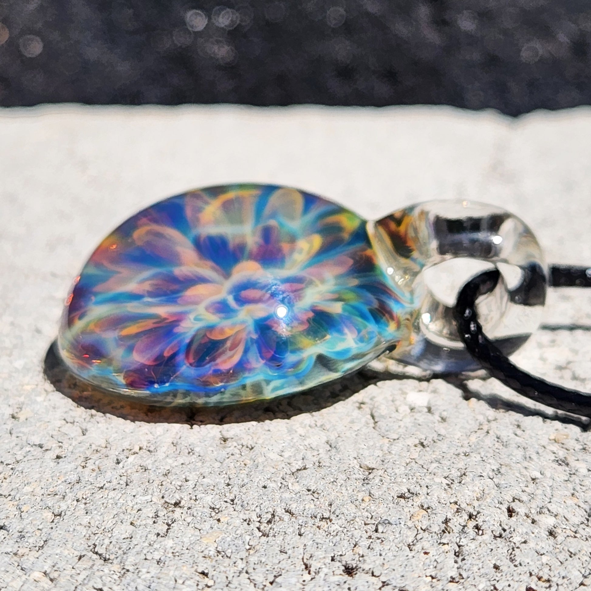Rainbow glass pendant with vibrant swirl pattern, on black cord, placed on white textured paper surface