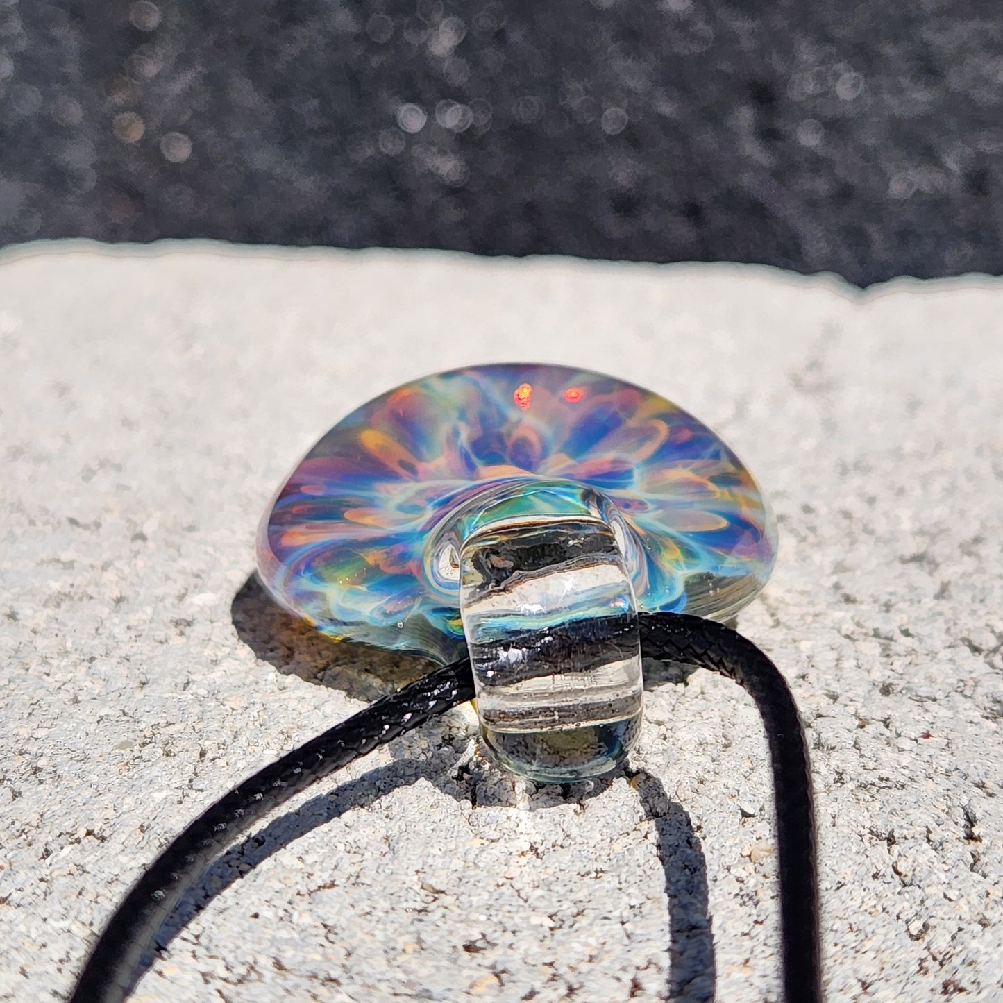 Colorful handmade glass pendant with swirling rainbow hues, teardrop shape, on a black braided cord, placed on a textured wooden surface, next to a quarter for scale.