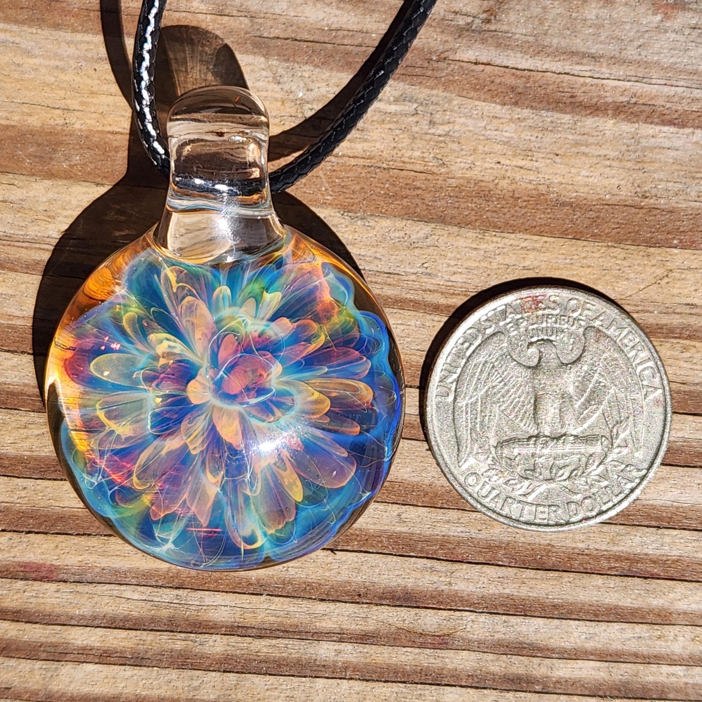 Close-up of the rainbow glass pendant's top with a clear glass loop, casting a shadow on the wooden background, alongside a quarter coin for size reference.