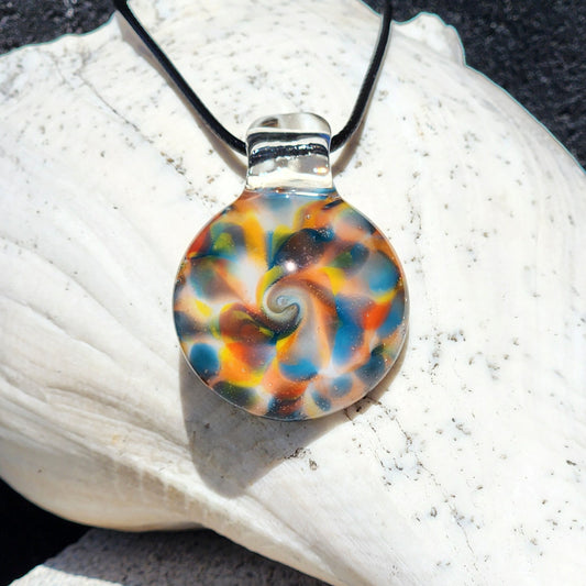 A round glass pendant necklace with a colorful spiral pattern in blue, orange, yellow, and brown, suspended from a black cord and resting on a white seashell.