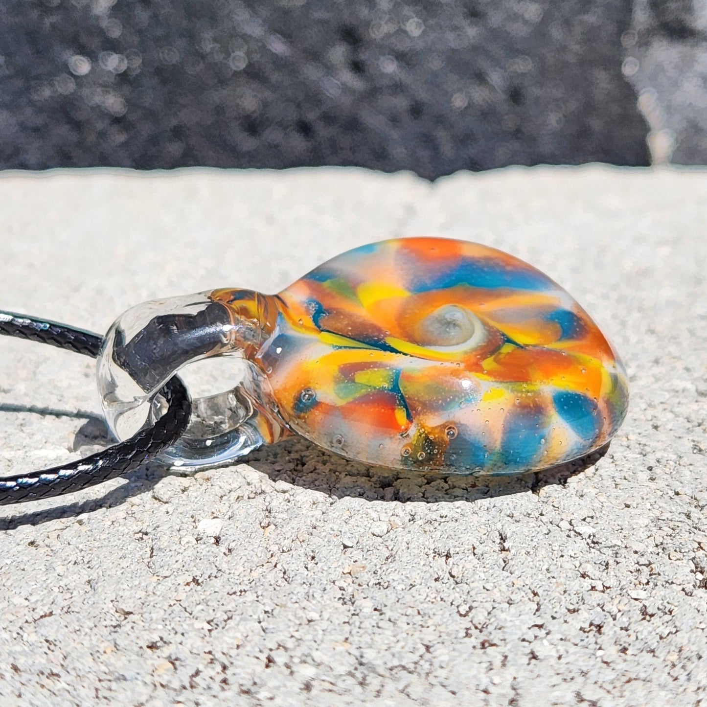 A glass pendant necklace with a vibrant swirl of orange, yellow, blue, and white colors, suspended on a black cord and resting on a textured concrete surface.