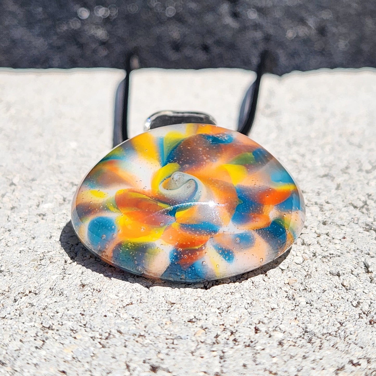 A colorful round glass pendant with a marbled swirl pattern of orange, blue, yellow, and white, resting on a light gray concrete surface outdoors.