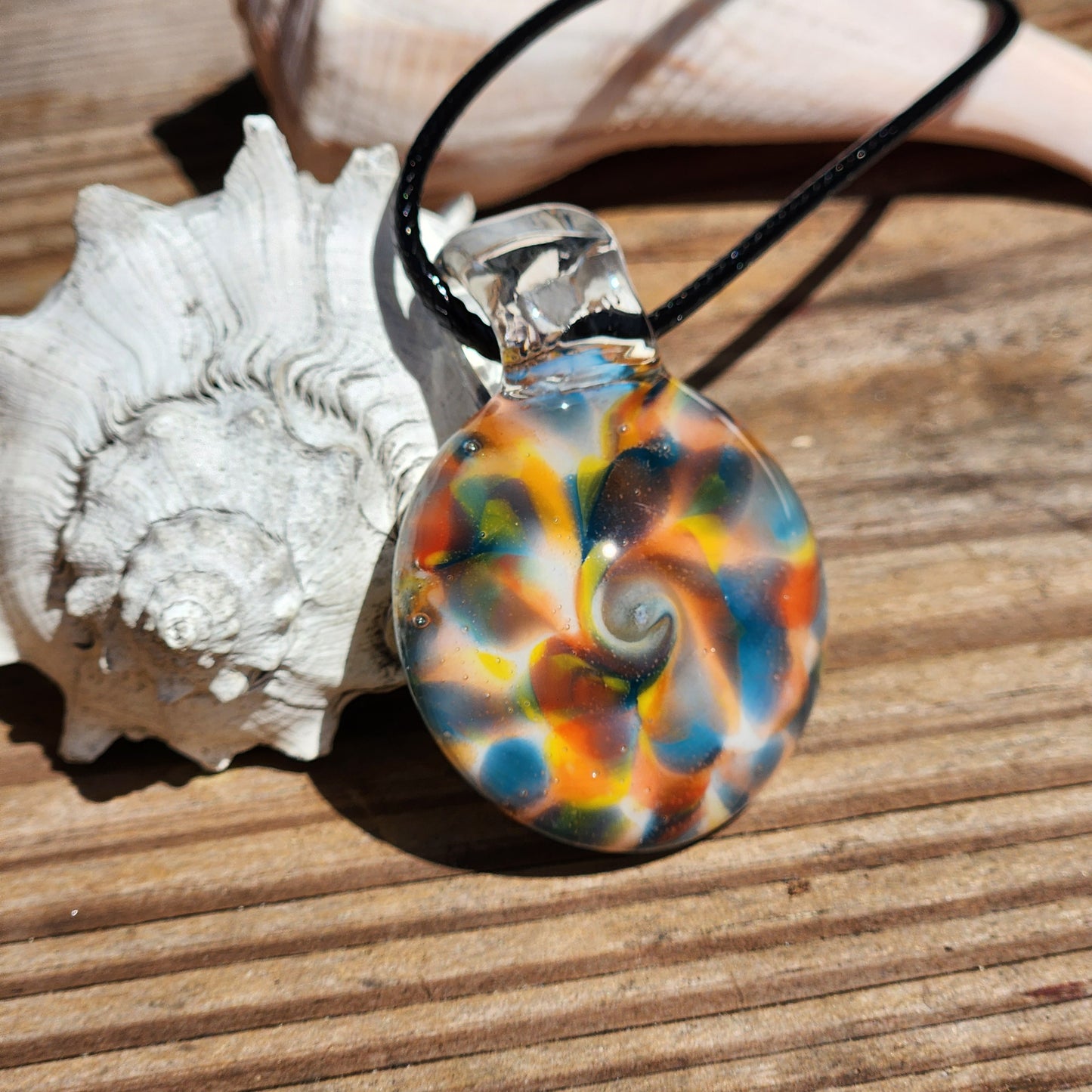 A round glass pendant necklace with a colorful seashell design, resting on a wooden surface next to a white seashell. The pendant features swirls of orange, blue, green, yellow, and white, suspended on a black cord.