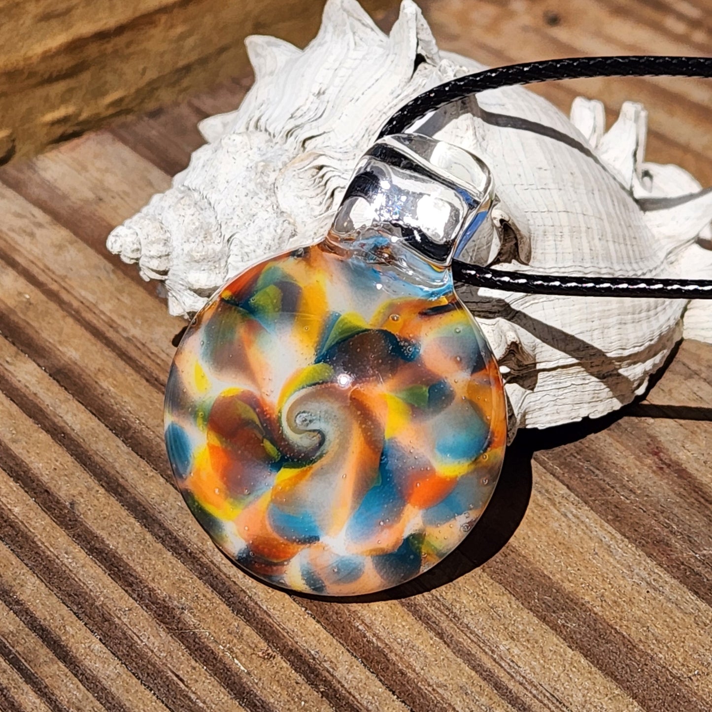 A round glass pendant necklace with a colorful seashell-inspired swirl pattern in blue, orange, yellow, and brown, resting on a white seashell over a wooden surface.