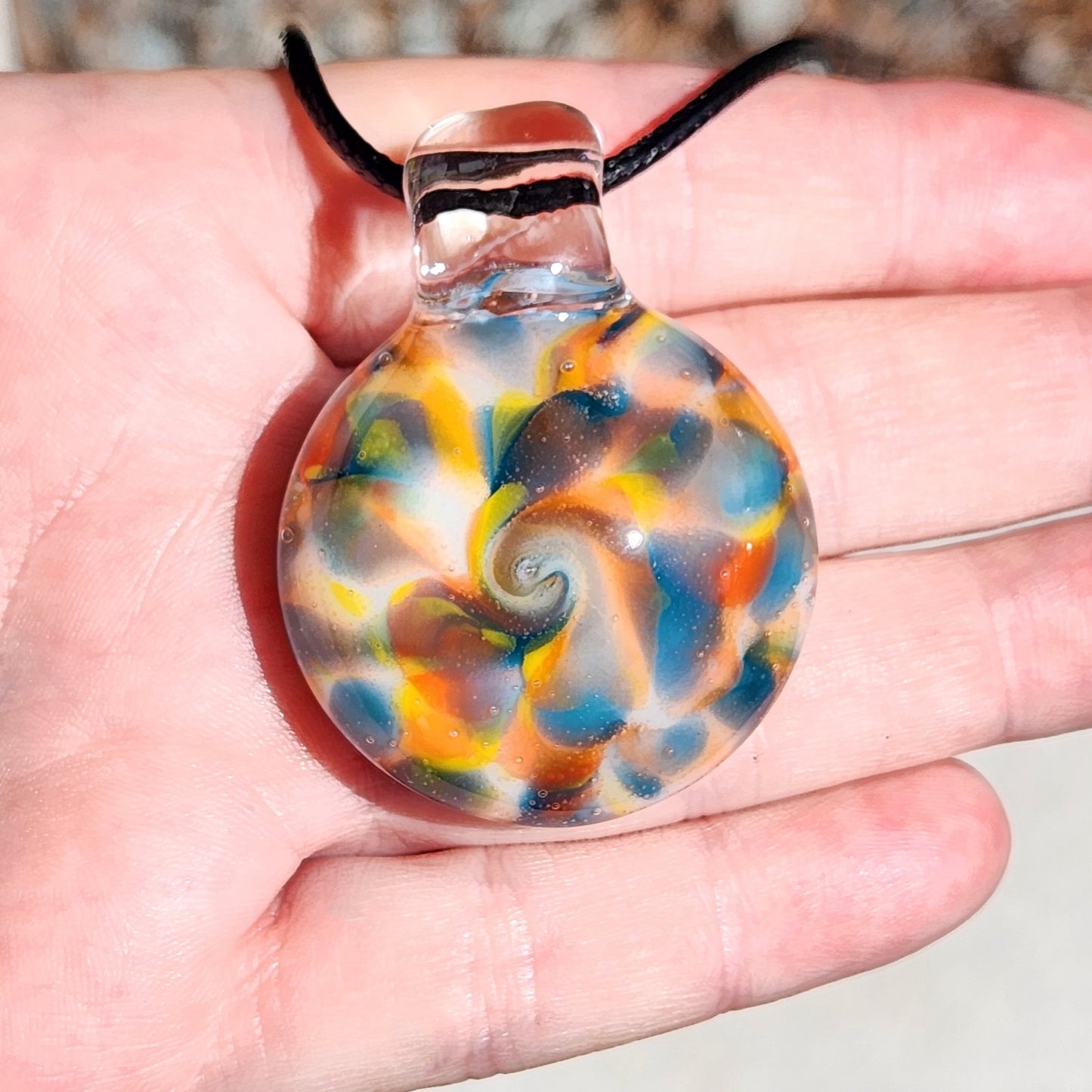 A hand holding a round glass pendant necklace with a colorful swirl pattern of blue, orange, yellow, and white, suspended on a black cord.