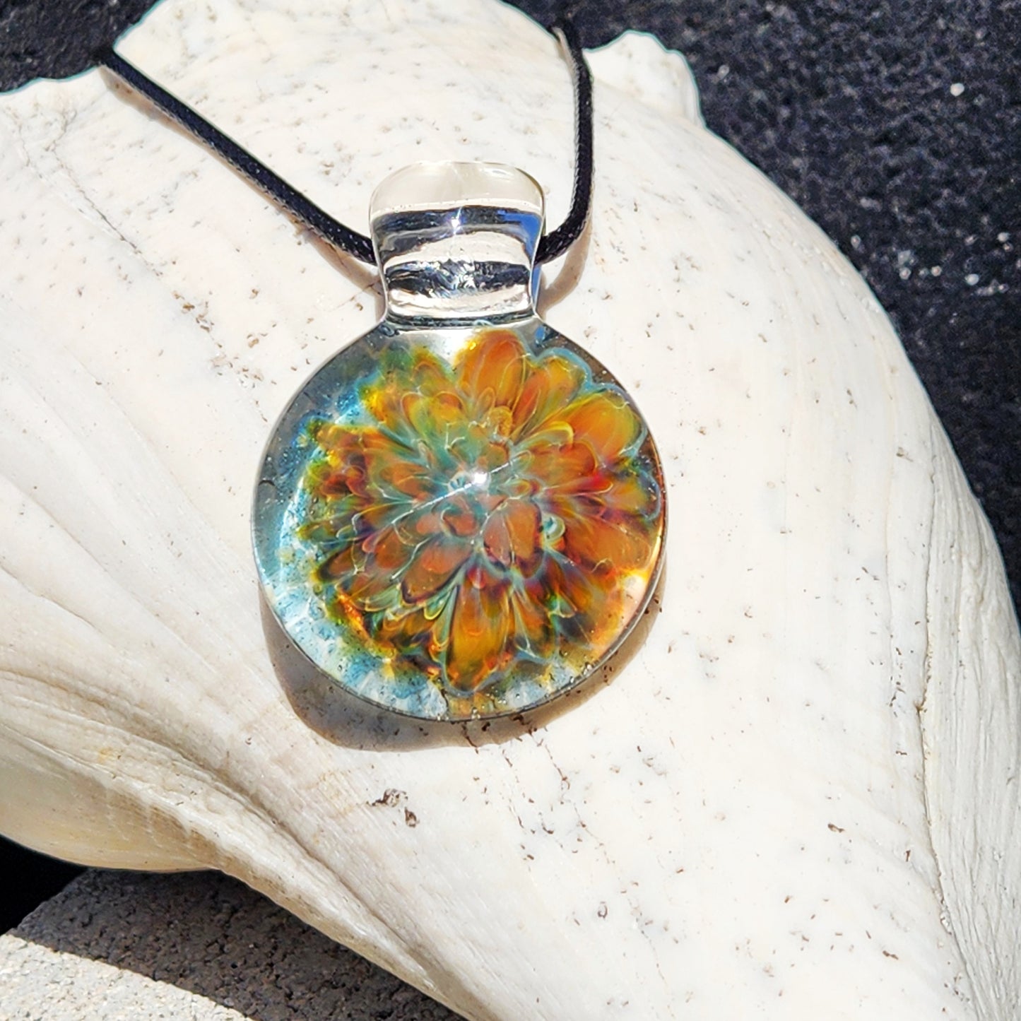 A round glass pendant necklace with a colorful floral mandala design in orange, yellow, and green, suspended from a black cord, resting on a large white seashell.