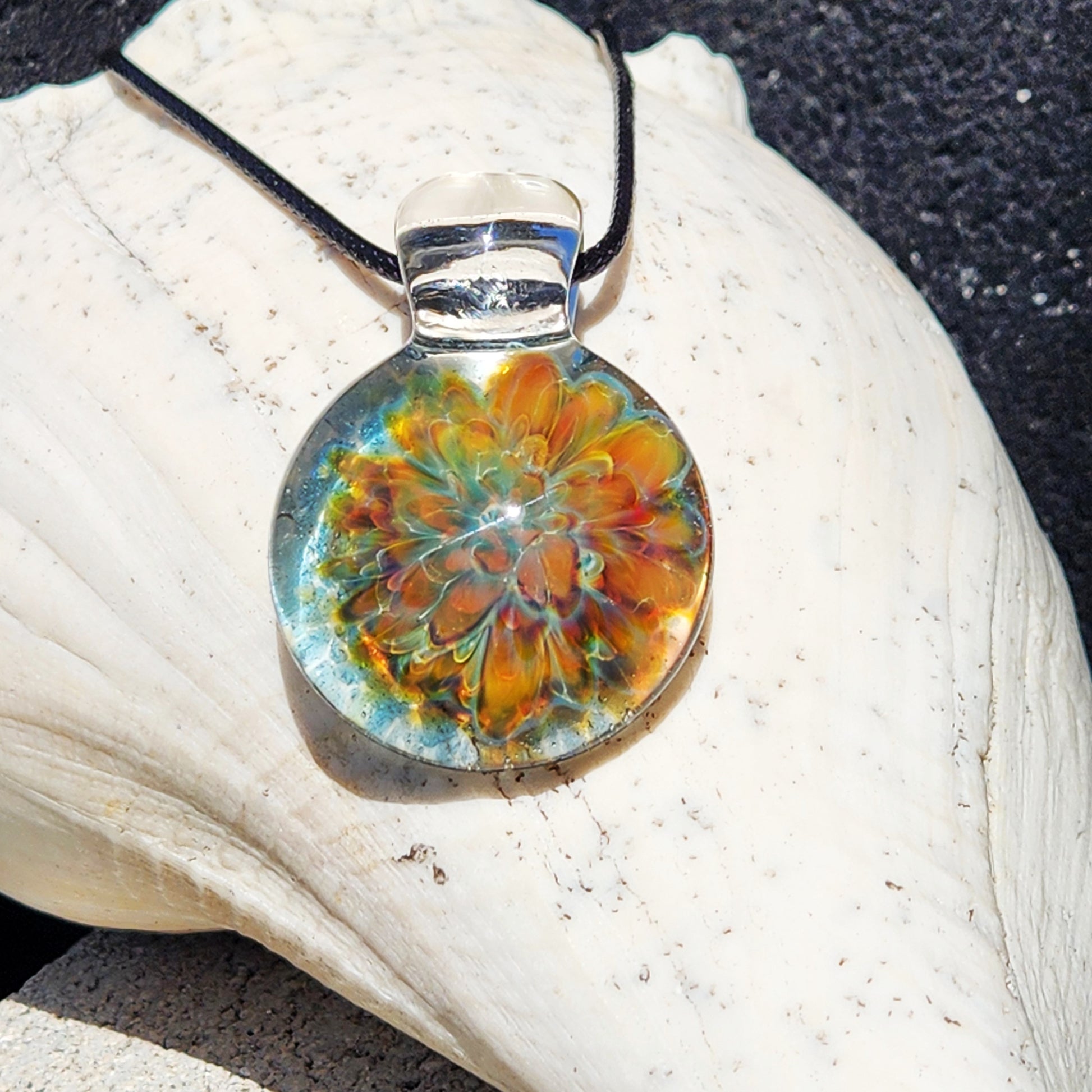 A round glass pendant necklace with a colorful floral mandala design in orange, yellow, and green, suspended from a black cord, resting on a large white seashell.