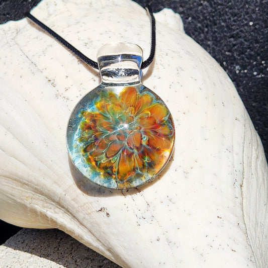 A round glass pendant necklace with a colorful floral mandala design in orange, yellow, and green, suspended from a black cord, resting on a large white seashell.