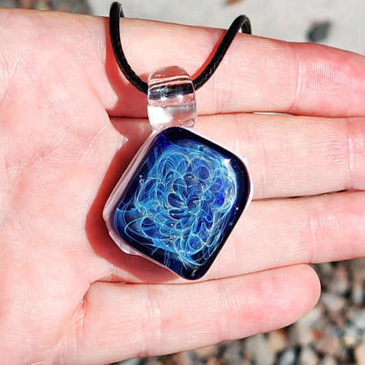 Close-up of a handmade glass pendant necklace with a deep blue swirling design, clear glass loop, and black cord, shown on a person's hand