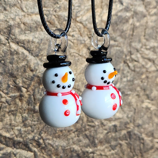 Two white snowman pendant necklaces with black hats, red scarves, and orange carrot noses hanging from black cords against a textured stone background.