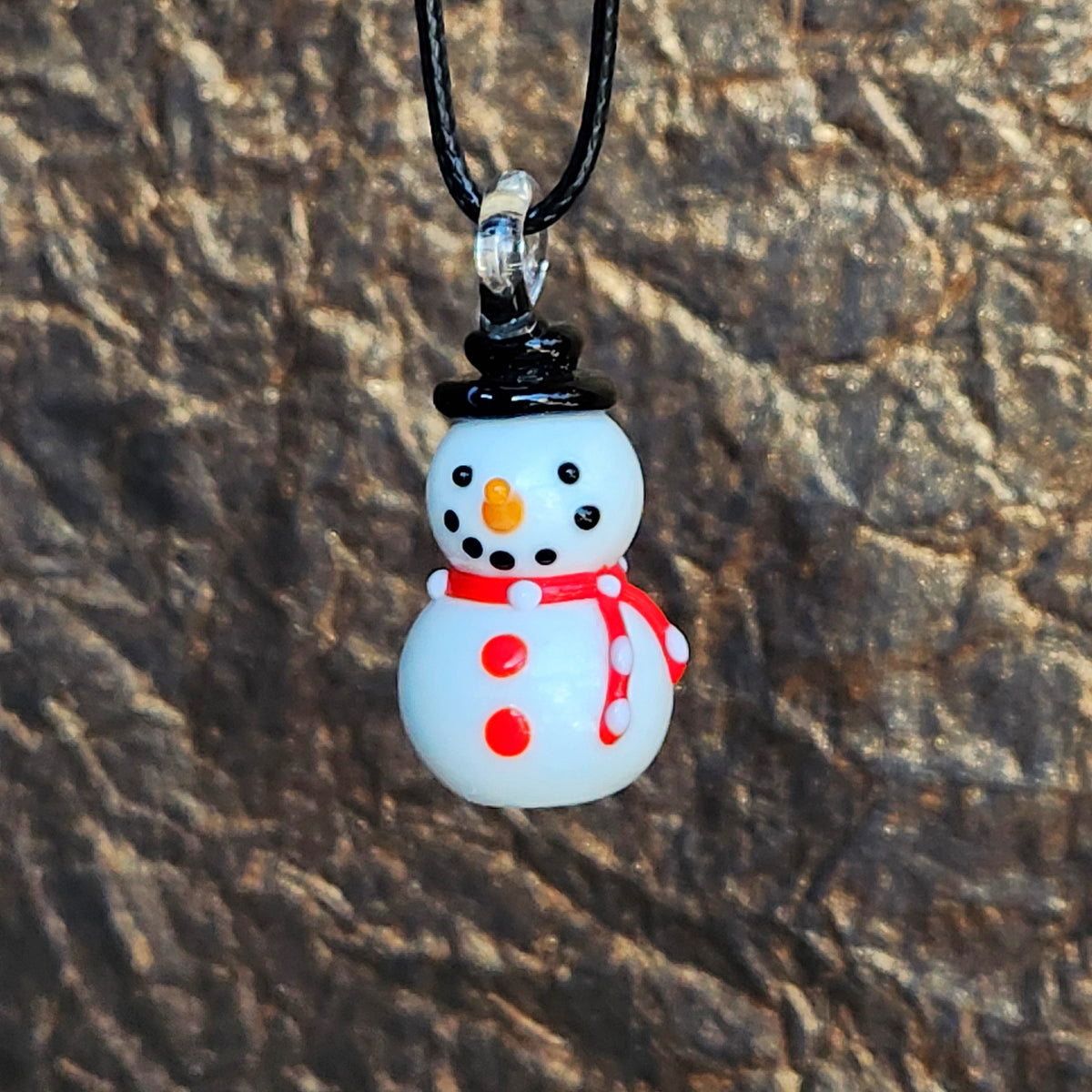 A small snowman pendant necklace with a black top hat, red scarf, and orange carrot nose, hanging from a black cord against a textured brown background.