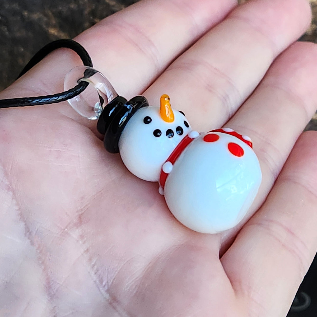 A close-up of a hand holding a small snowman pendant necklace. The pendant is made of glossy white glass with a black hat, orange carrot nose, black dot eyes, and a red-and-white polka-dot scarf, attached to a black cord.