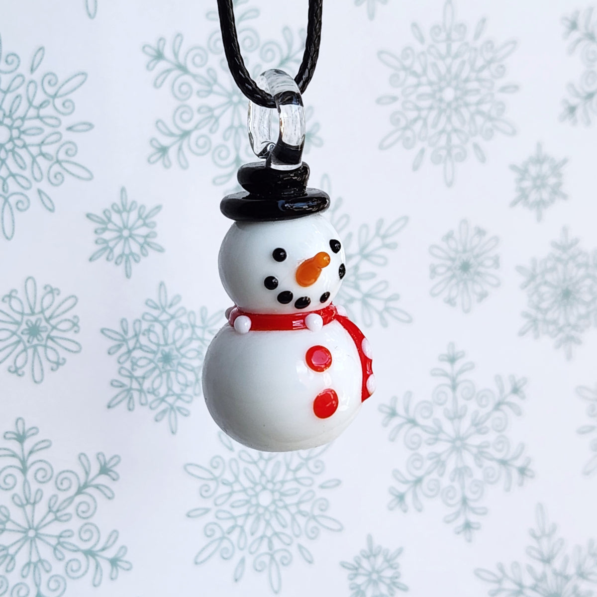 A small snowman pendant necklace with a black cord, featuring a white body, black hat and buttons, a red scarf, and an orange carrot nose, set against a white background with a light blue snowflake pattern.