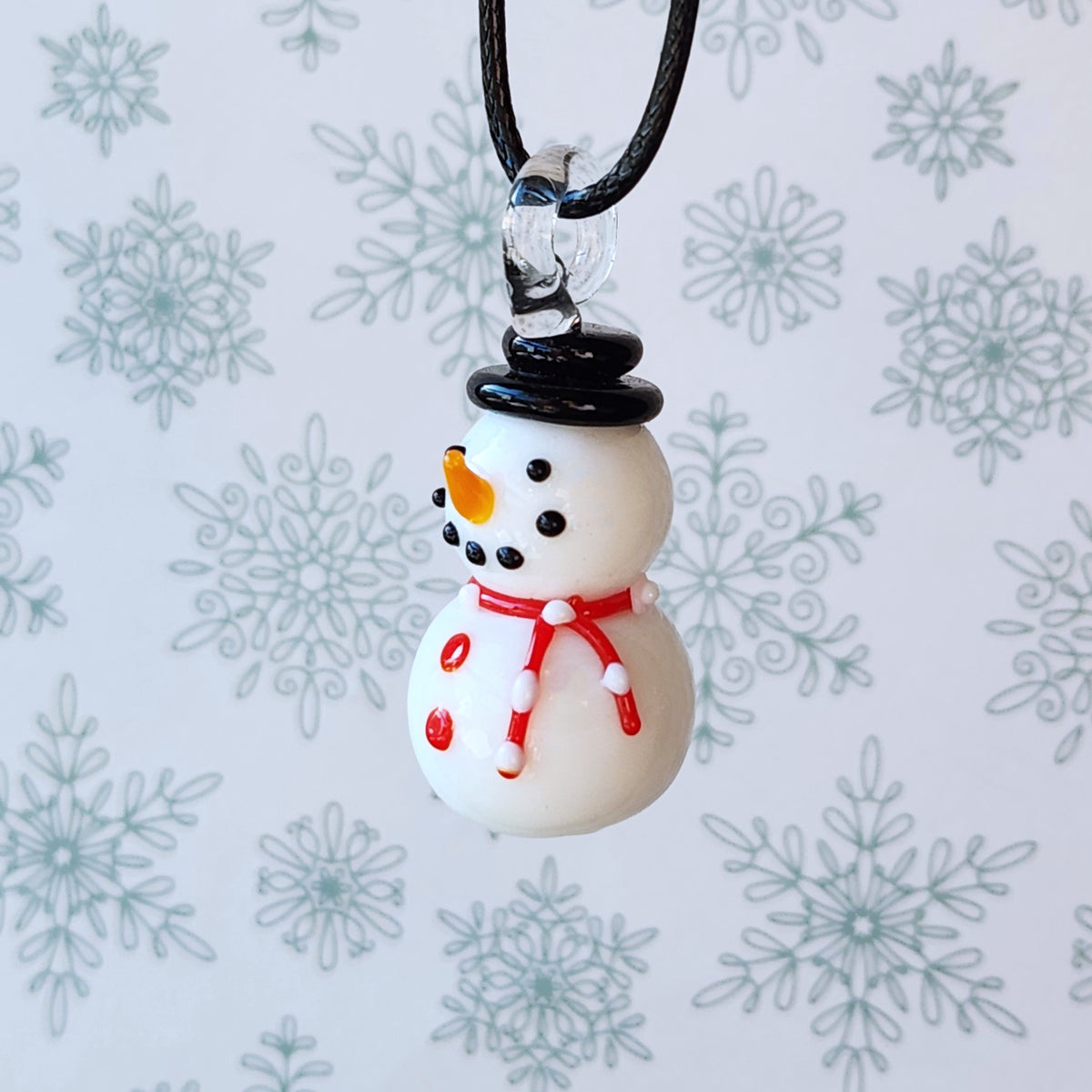 A small snowman pendant necklace with a black cord, featuring a white body, black hat and buttons, an orange carrot nose, and a red scarf, shown against a light background with gray snowflake patterns.