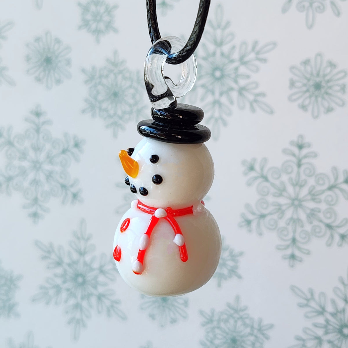 A small snowman pendant necklace with a black top hat, red scarf, and orange carrot nose, hanging from a black cord against a white background with a blurred snowflake pattern.
