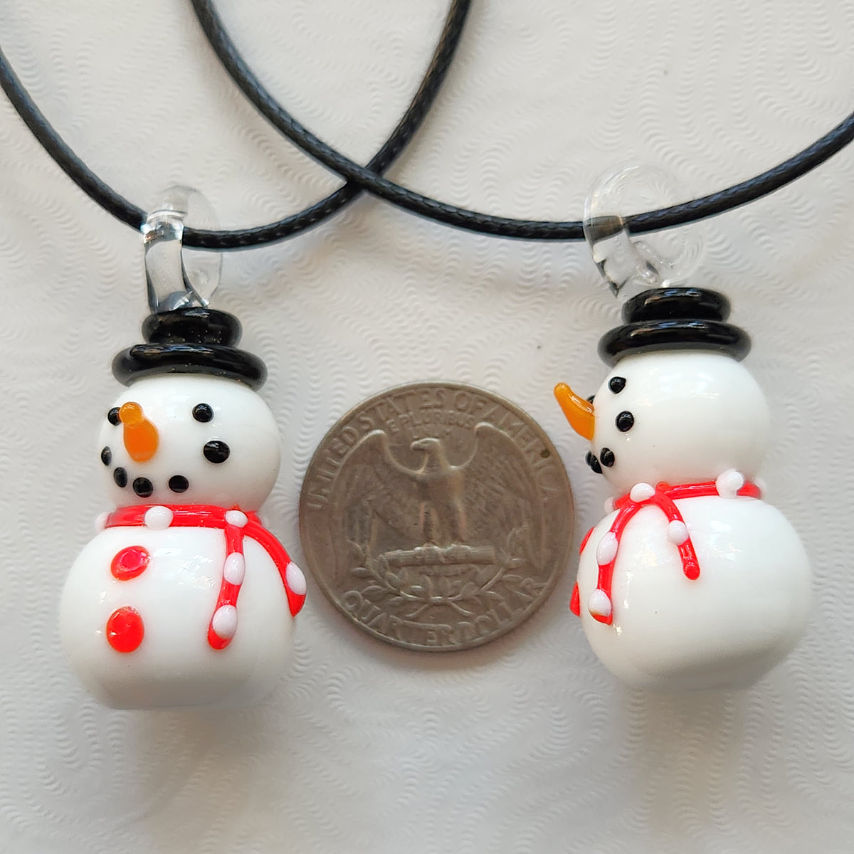 Two white snowman pendant necklaces on black cords, shown next to a US quarter coin for scale on a textured white background.